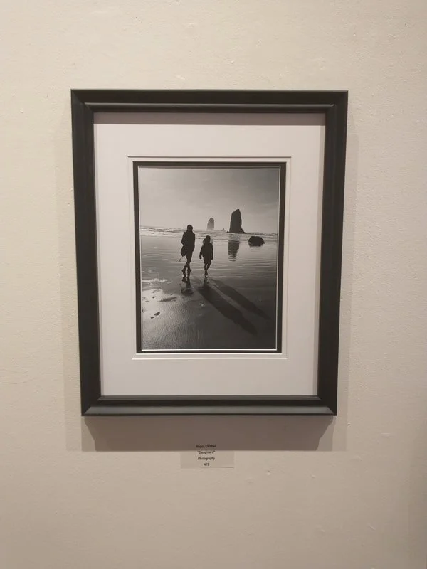 Black and white photograph of two people walking on a beach with large rock formations in the background, casting long shadows on the wet sand.