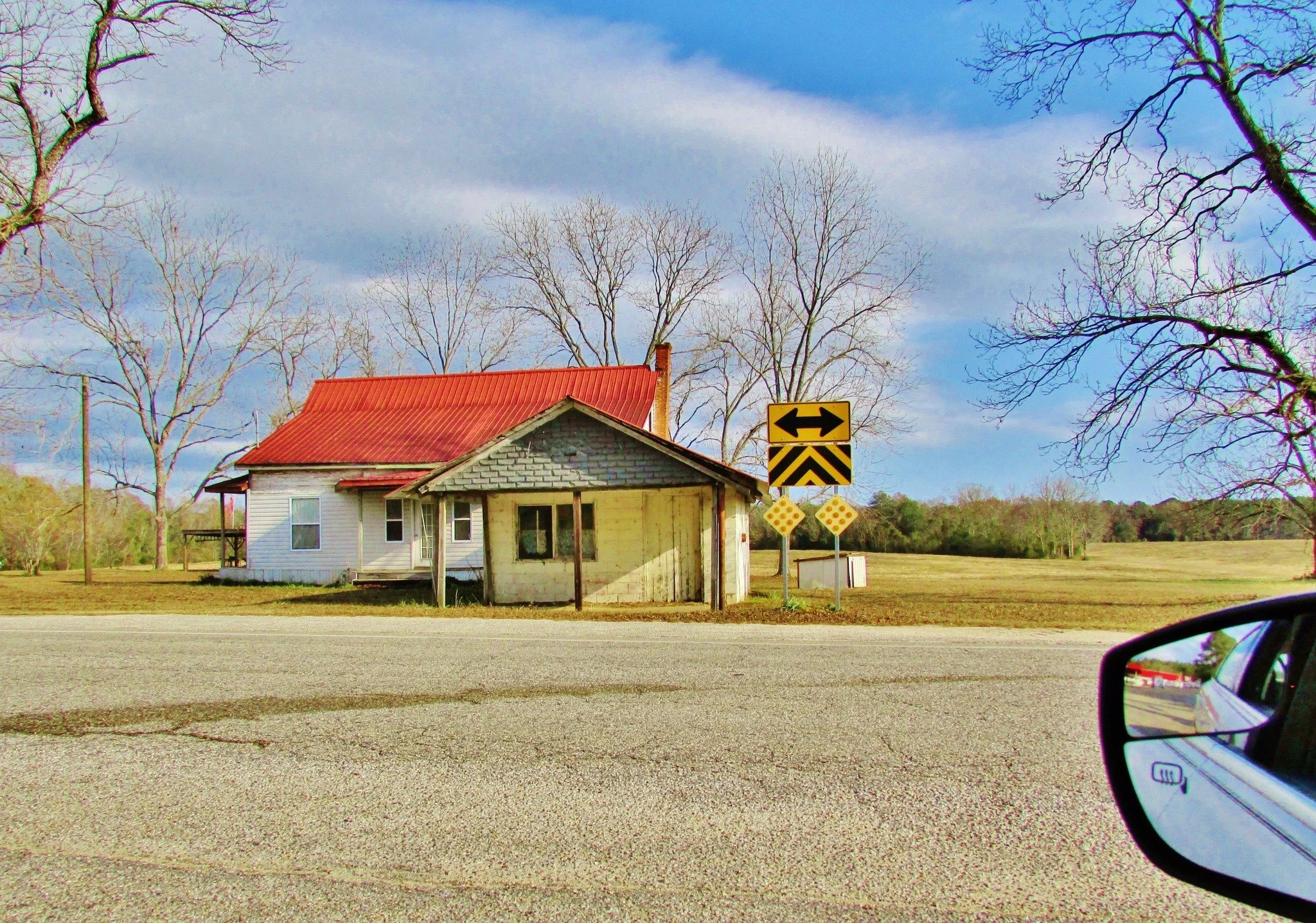 A house with a red metal roof and yellow brick exterior, surrounded by leafless trees, under a partly cloudy sky, with a road in the foreground and road signs nearby.
