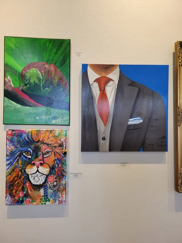 Three colorful paintings on a wall: a green abstract landscape with a hobbit-like figure, a vibrant lion's head portrait, and a grayscale portrait of a man in a suit with a red tie.