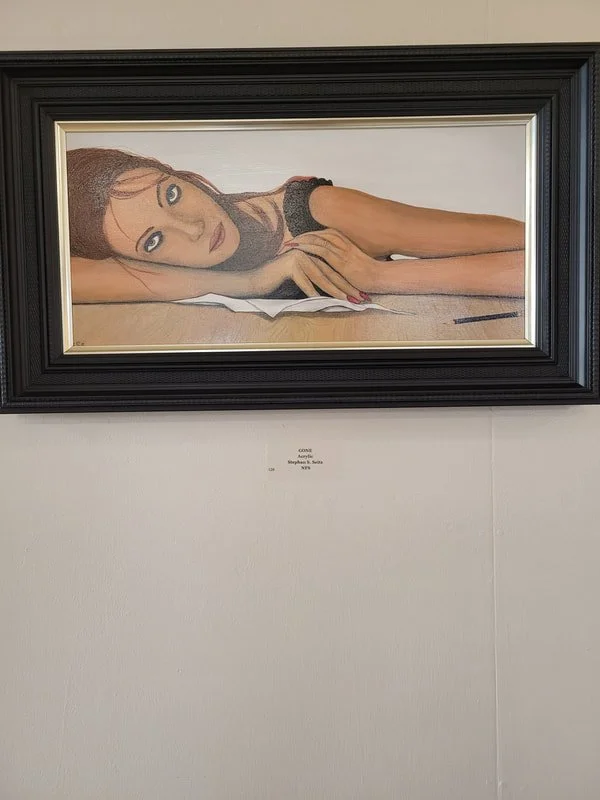 Framed painting of a woman lying on a wooden surface with her head resting on her hands, wearing a black top, with brown hair and light skin.