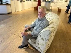An elderly man with white hair sitting on a large floral-patterned armchair in a store, holding a remote control, smiling at the camera.