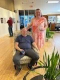 A woman standing beside a man who is seated in a wheelchair in an indoor setting, with other people in the background and potted plants nearby.