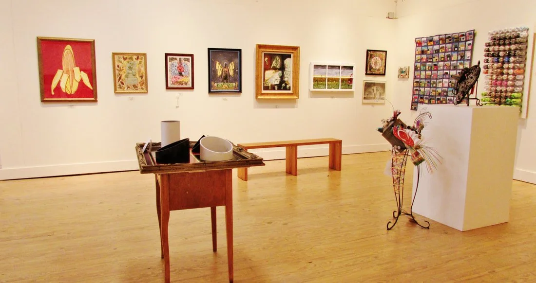 An art gallery with framed paintings and artworks on white walls, a wooden table with art supplies, a wooden bench, and a sculpture with flowers and ribbons.