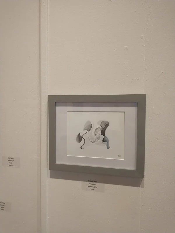 A framed abstract artwork depicting multiple stylized, organic shapes in shades of gray and black, hung on a white gallery wall.