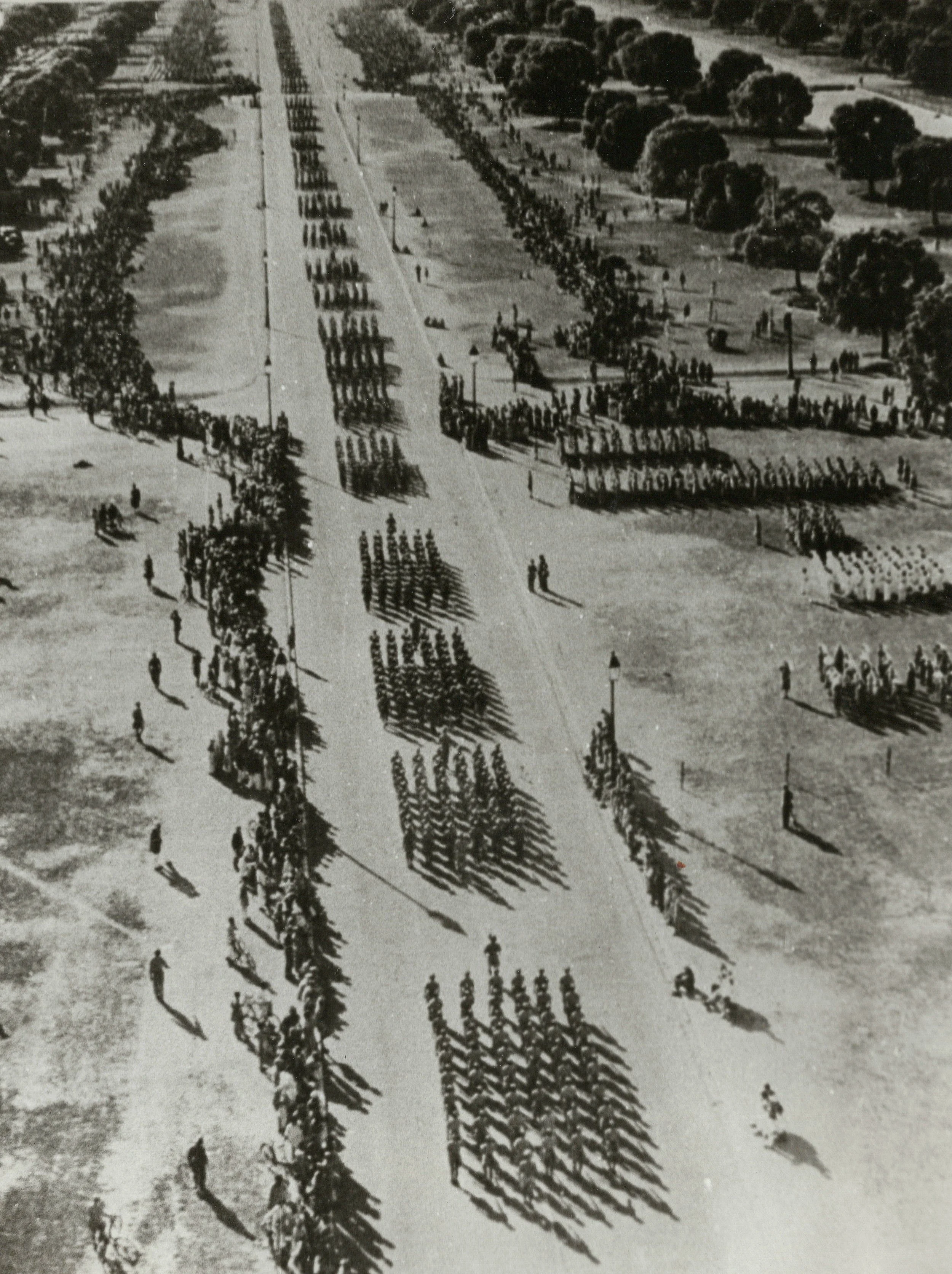An aerial black and white photograph of a large gathering of troops, organized formation with rows of windbreaks, and a long central pathway lined with street lamps and trees.