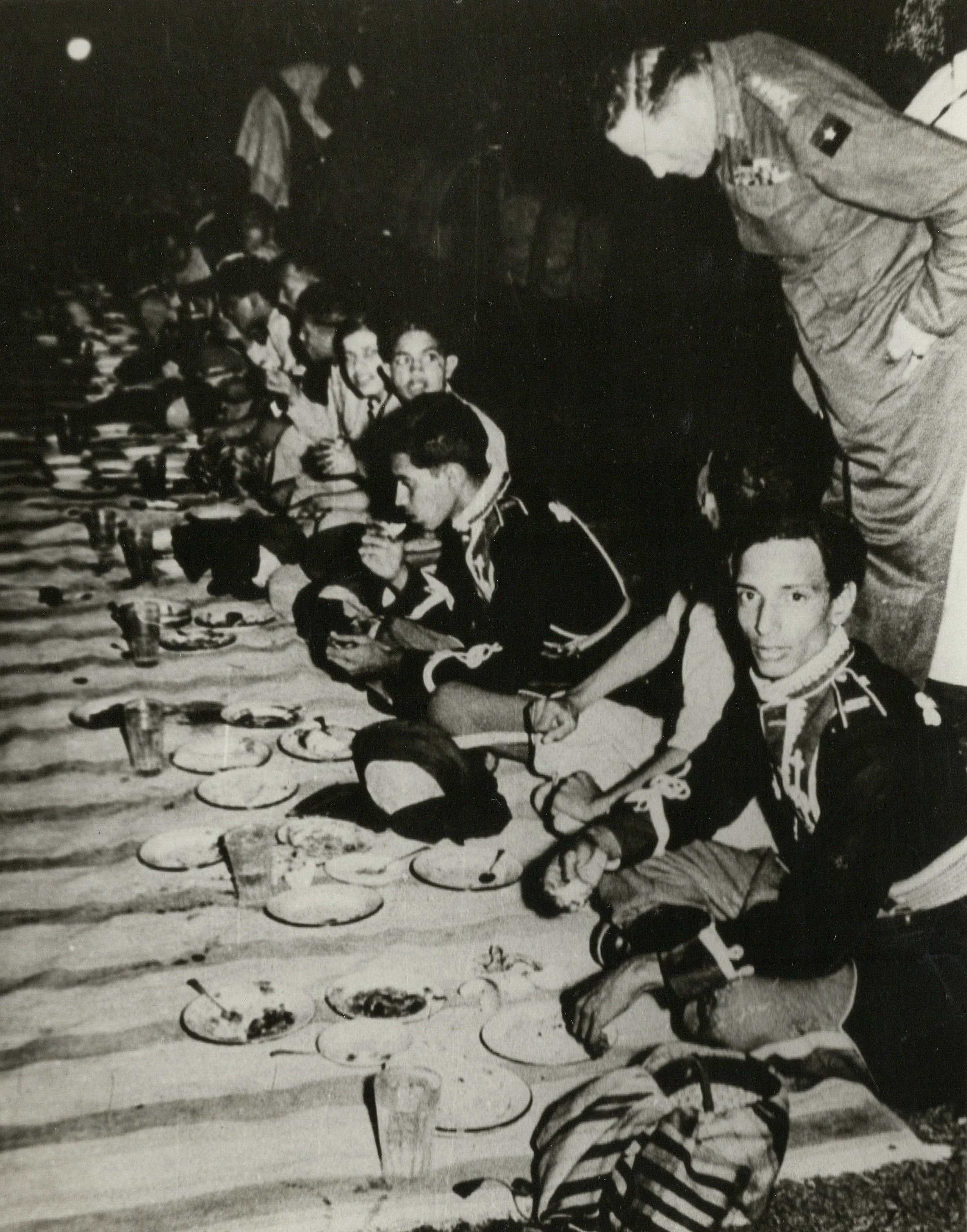 People sitting on a striped blanket at a picnic with plates of food, cups, and drinks arranged in front of them, with one person in the foreground wearing a striped sweater and others in the background, some wearing uniforms, suggesting a gathering or celebration.