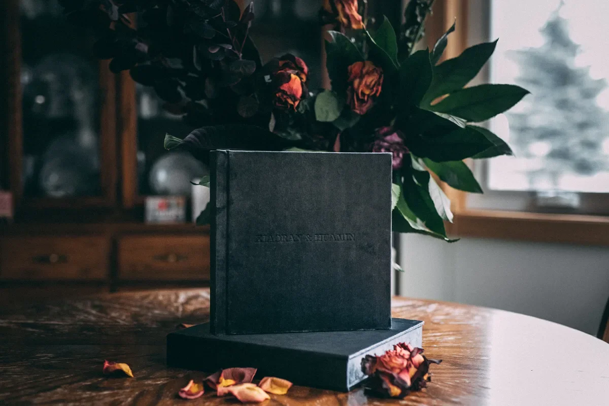Navy velvet wedding album in a vertical flat lay with rose petals and green leaves-thin-bordered