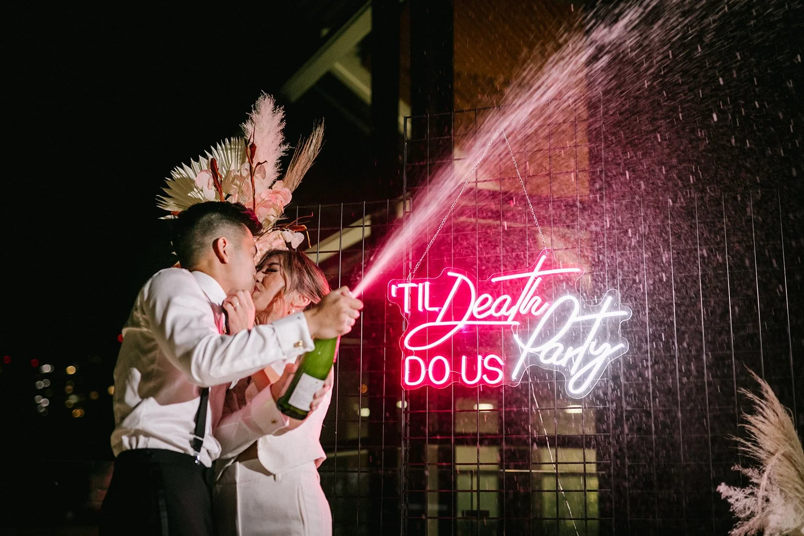 A kissing wedding couple pops a bottle of champagne in front of a neon sign that says "'til death do us party"
