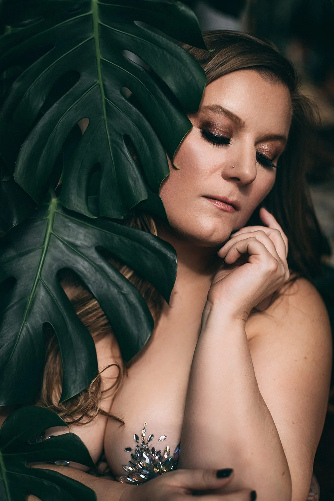 a woman wearing crystal nipple pasties poses beside a large monstera leaf