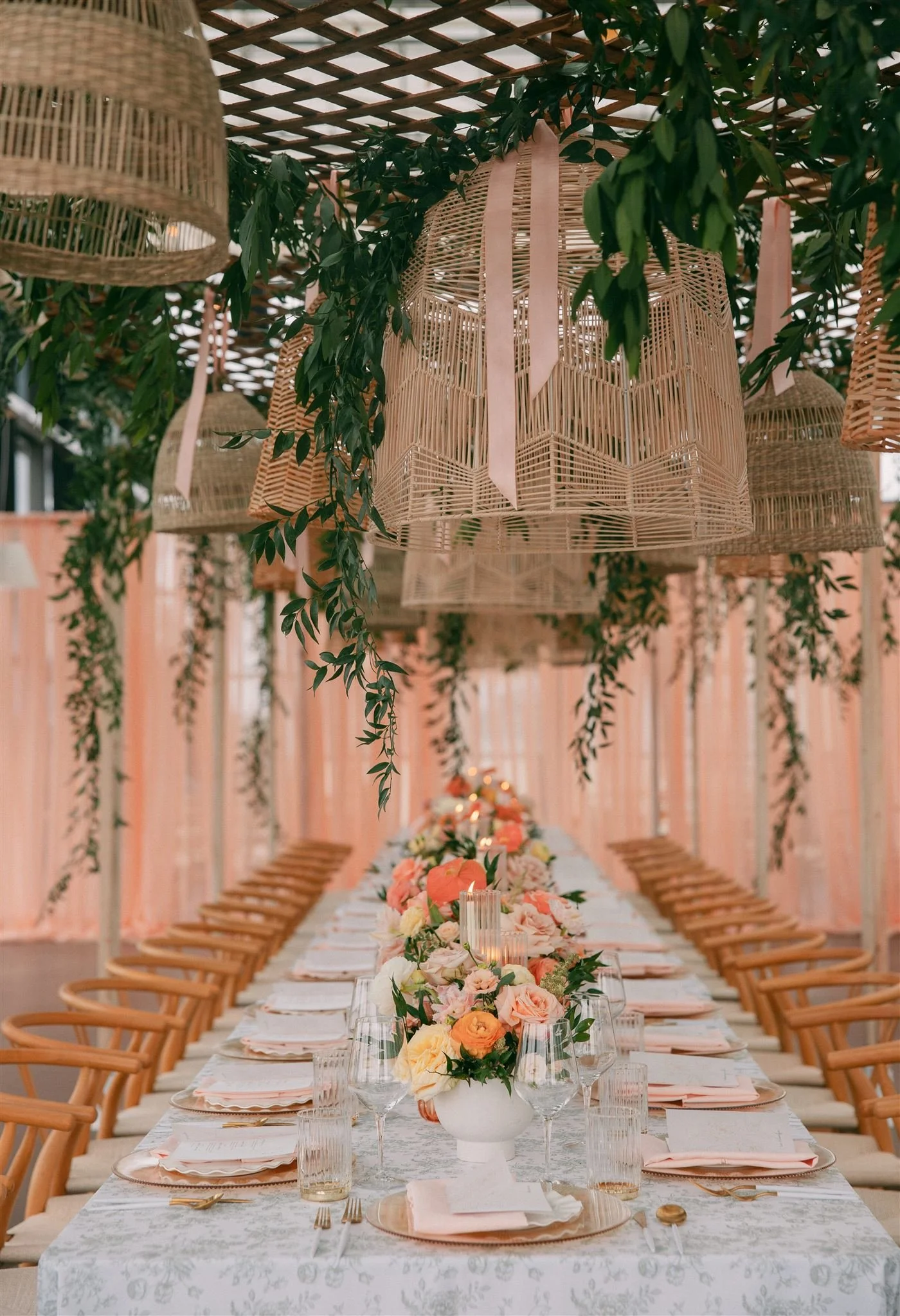 a long king's style tablescape set for a brunch wedding reception, with wicker lanters and greenery hanging down overhead and bright peach florals on the table