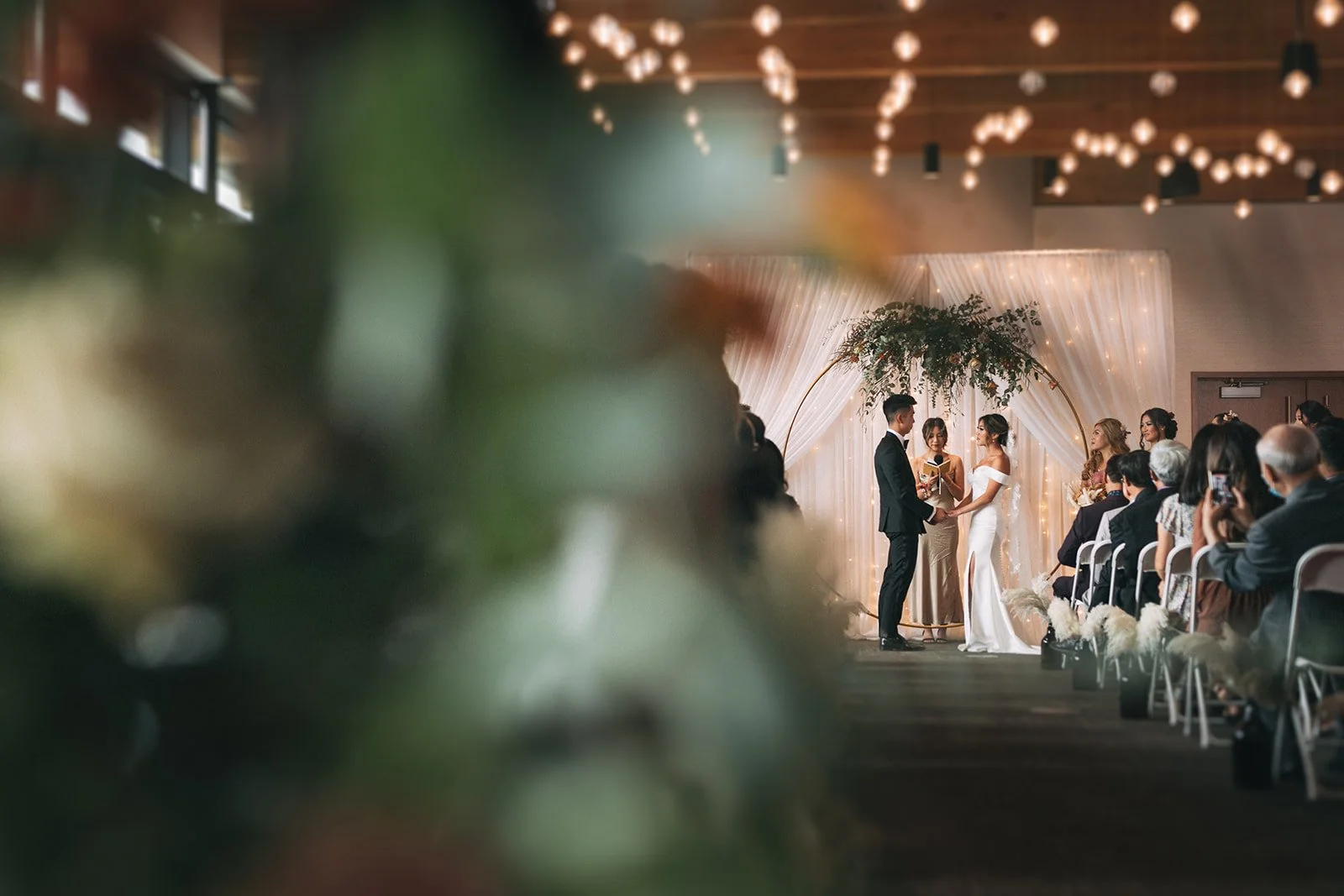 A couple stands under a circular altar holding hands during a wedding ceremony at the Royal Glenora Club. Foliage partially obscures the frame and lights twinkle overhead