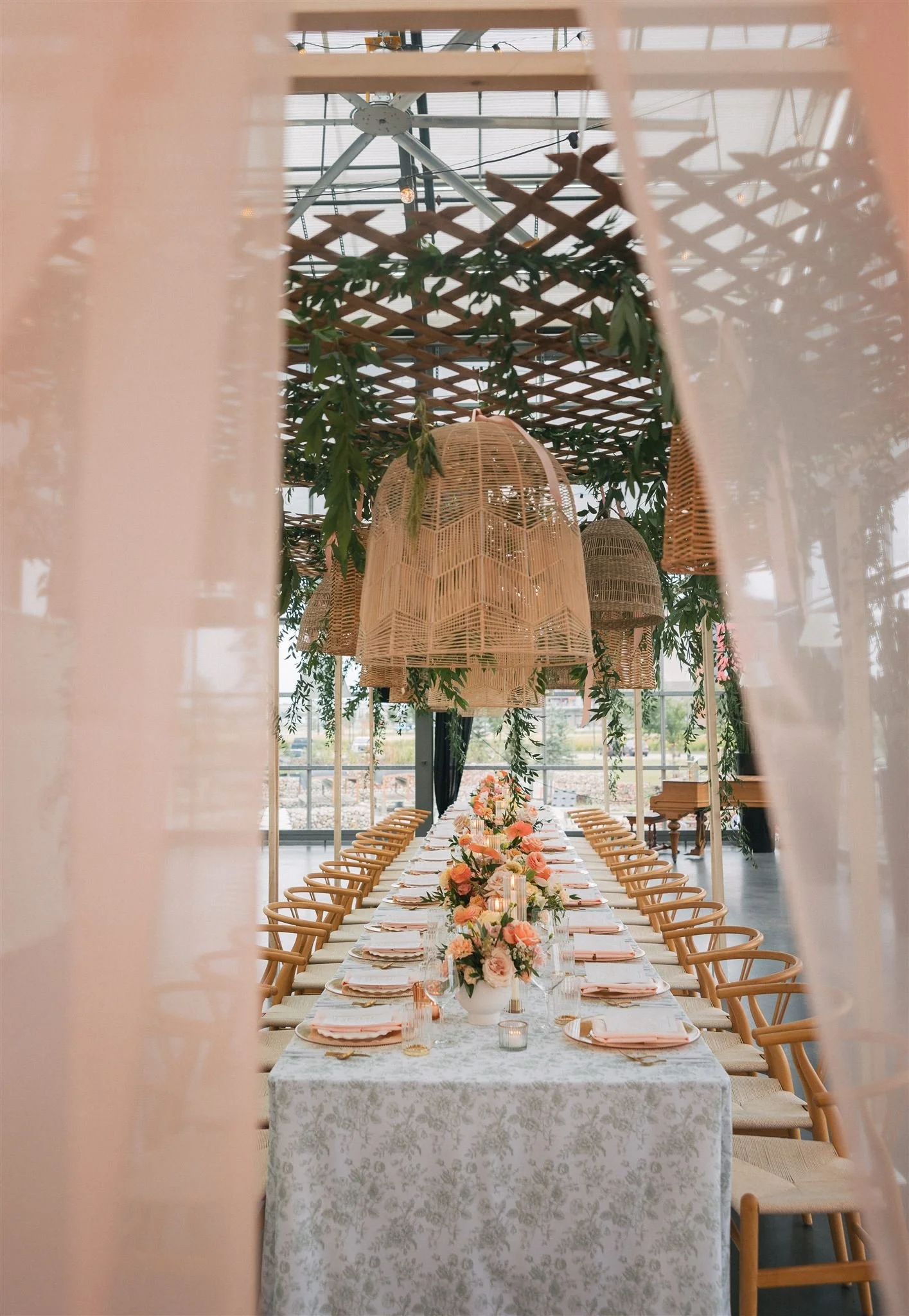 peeking through gauzy peach curtains, there is a wedding table with toile tablecloths, peach florals, and wicker lanters hanging overhead