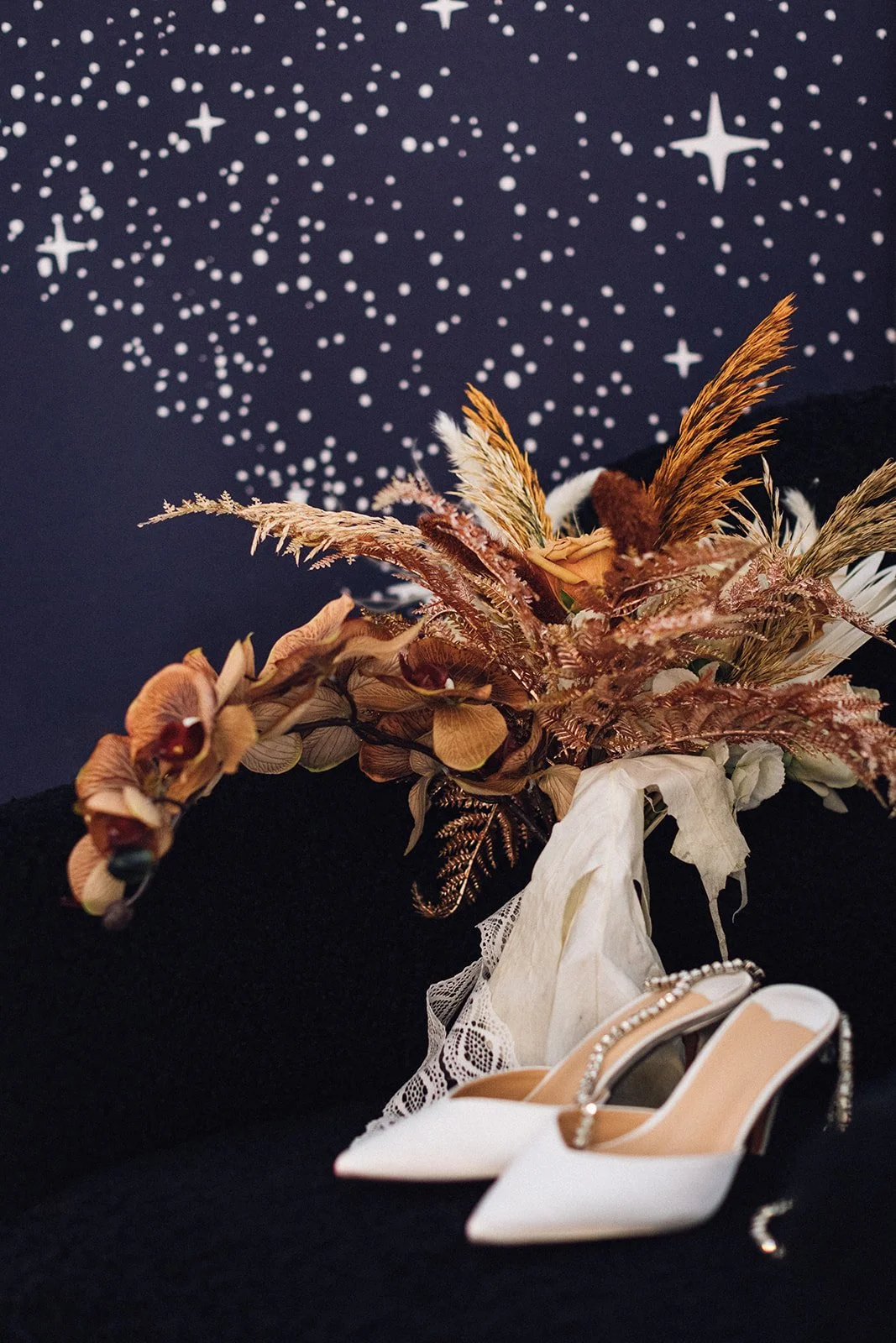 an autumn-themed wedding bouquet waits with a pair of white bejewelled heels, with a painting of stars in the background