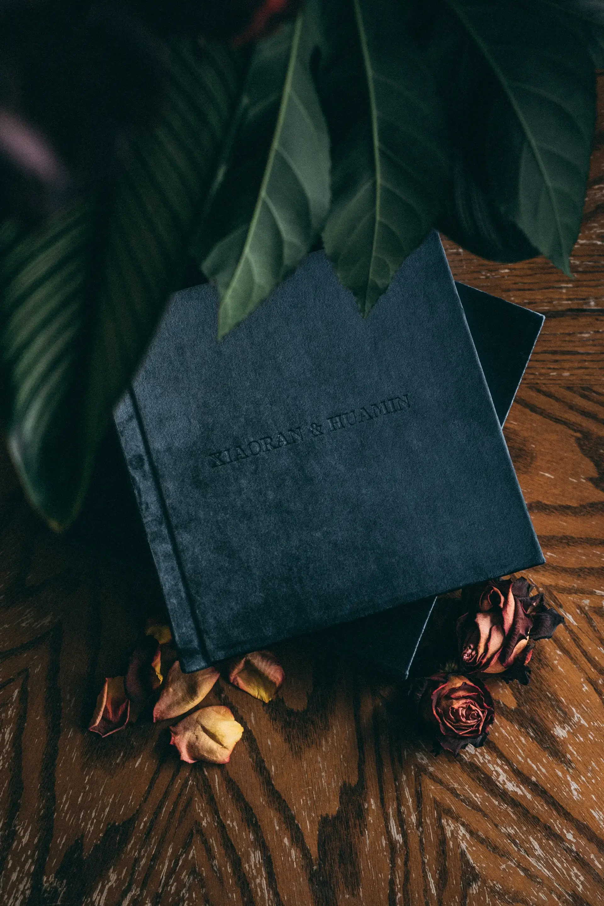 Navy velvet wedding photo album lying flat surrounded by dried roses and greenery