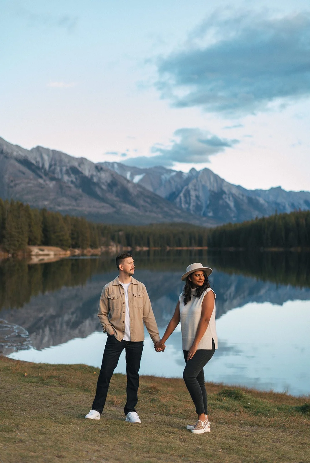 A man wearing a tan jacket and black jeans holds the hand of his fiancee, wearing a wide-brimmed hat and soft mock neck against the background of Johnson Lake, Banff
