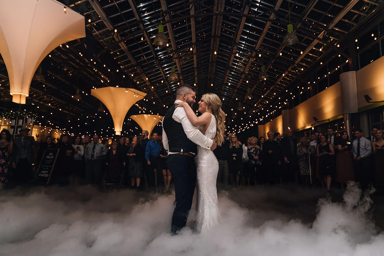 A couple shares their first dance with guests watching in the background, twinkle lights overhead, and fog rolling around their feet