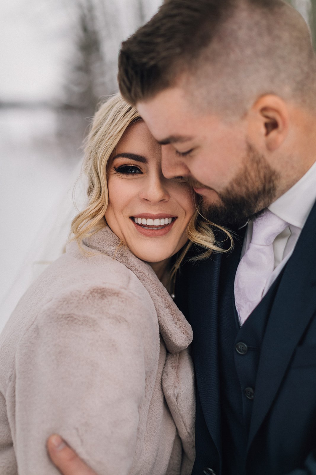 A groom keeps his bride warm on their winter wedding day, she peeks out at the camera with one blue eye