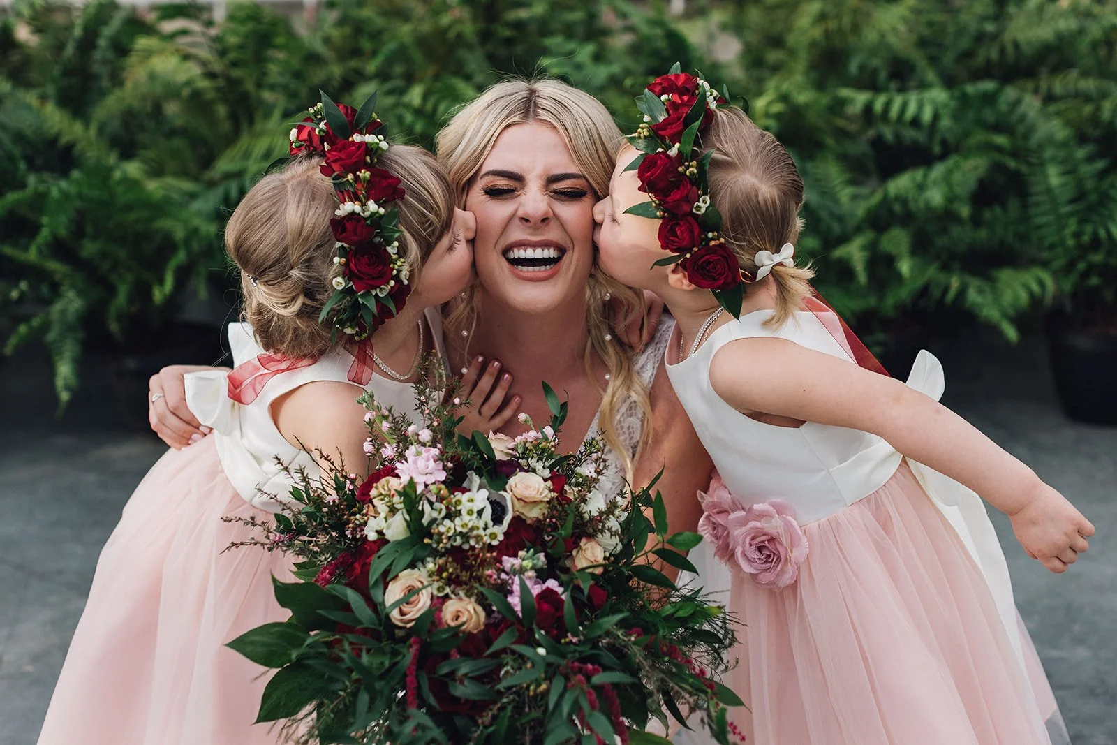 two flower girls wearing red rose flower crowns kiss the bride on the cheeks while she laughs
