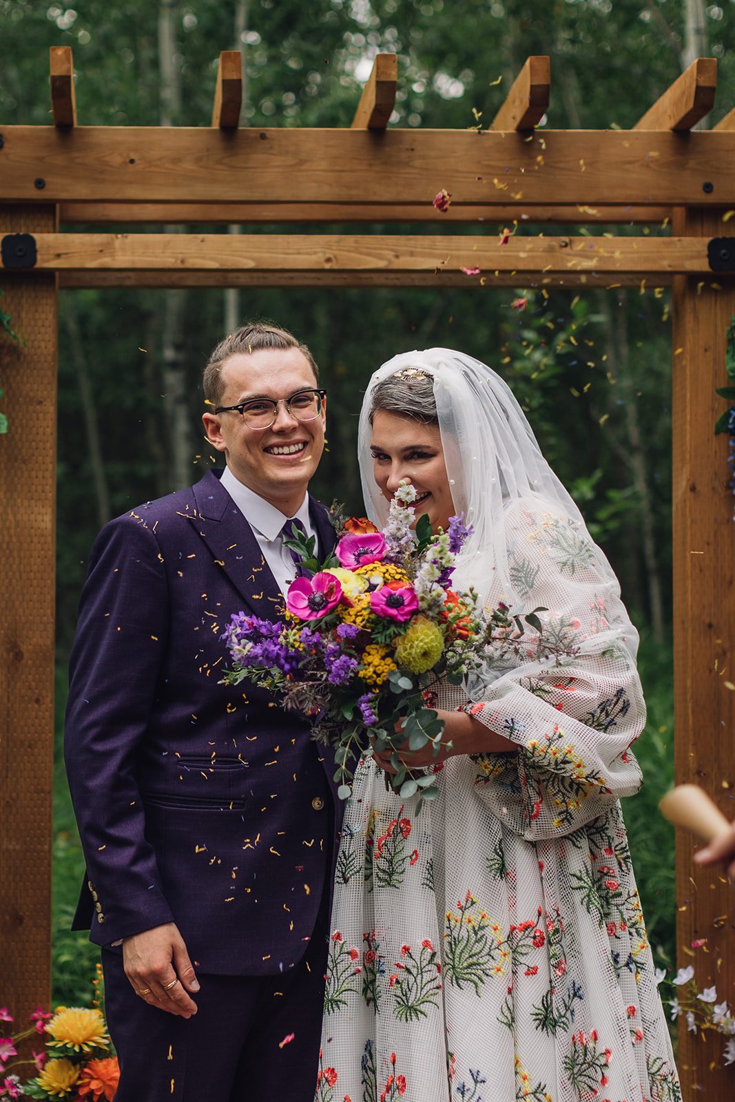 a colourful wedding portrait of a groom in a purple suit and his bride standing under an arch with flower confetti