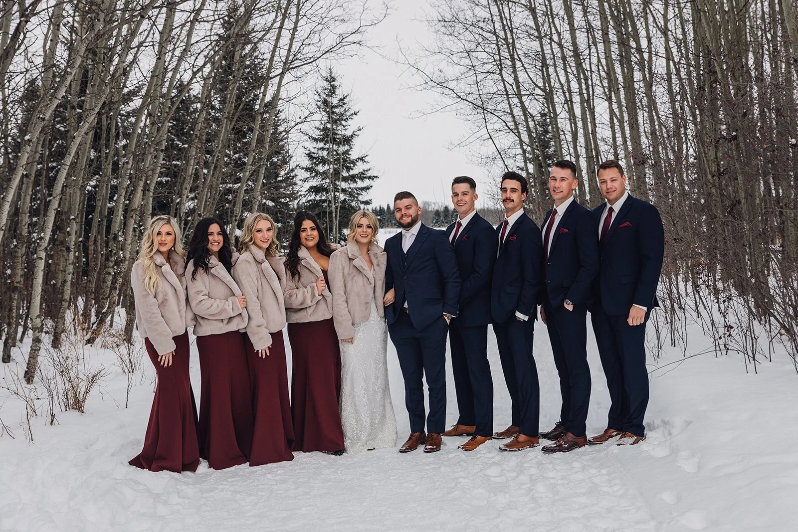 a wedding party poses for a photo, the bridesmaids are in burgundy dresses with pink faux fur coats and the groomsmen are in navy suits