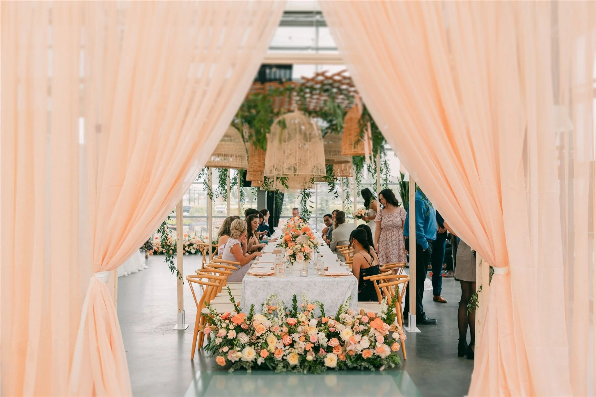A long wedding table peeks through peach coloured curtains showing wedding guests mingle over brunch while wicker lanterns and greenery hang overhead