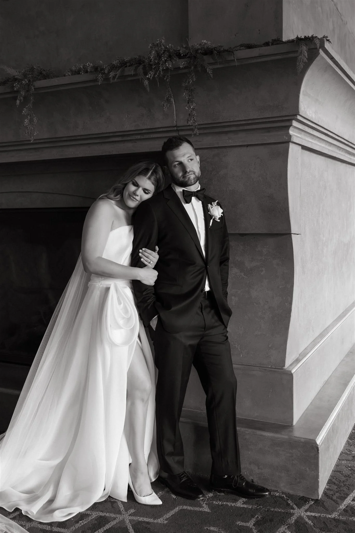 wedding portrait of a bride and groom standing by a large fireplace and she's resting her head on his shoulder-thin-bordered