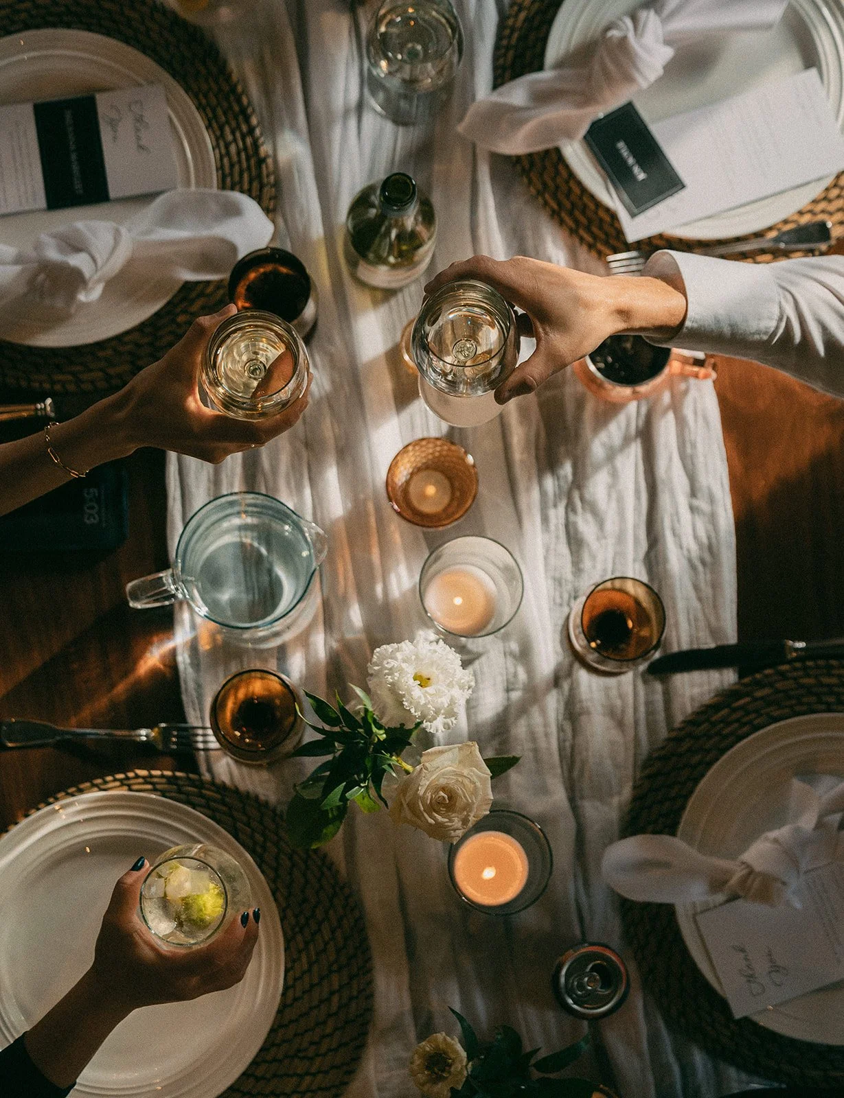 overhead view of a styled wedding table with candles, flowers, place settings, and two hands cheersing-thin-bordered