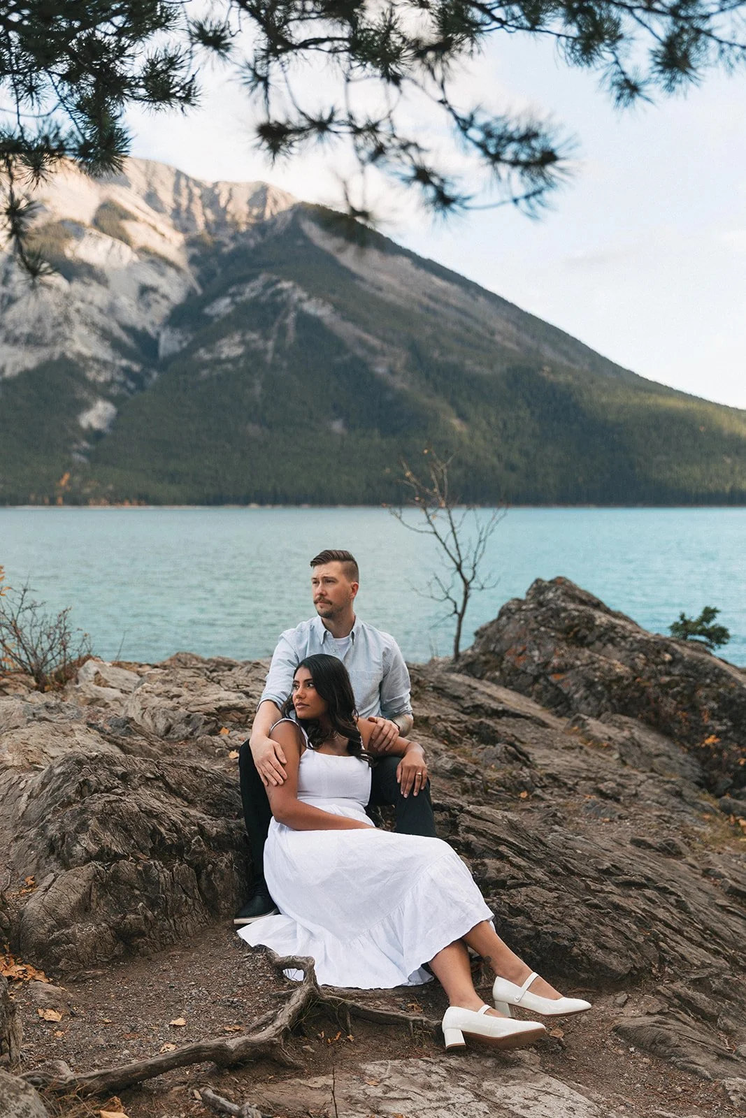 An engaged couple sits on rocks together and looks out at Lake Minnewanka, with Mount Inglismaldie in the background