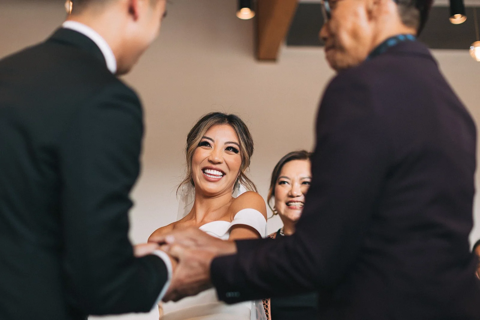 A bride smiles excitedly at her groom as she's walked to the front of the aisle. You can see her father holding her groom's hands in welcome in the foreground