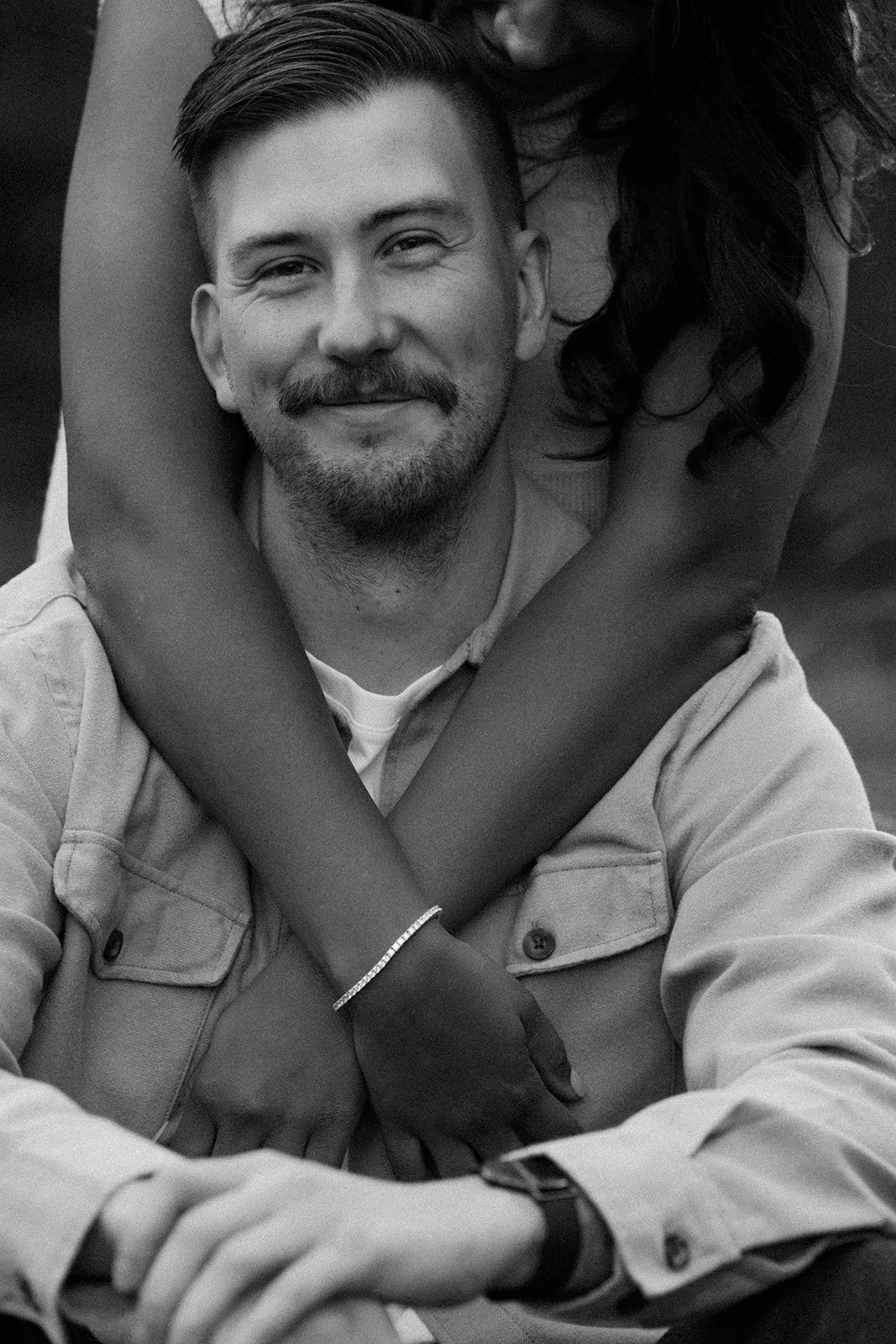 A black and white image of a man smiling softly at the camera while his fiancee hugs him from behind