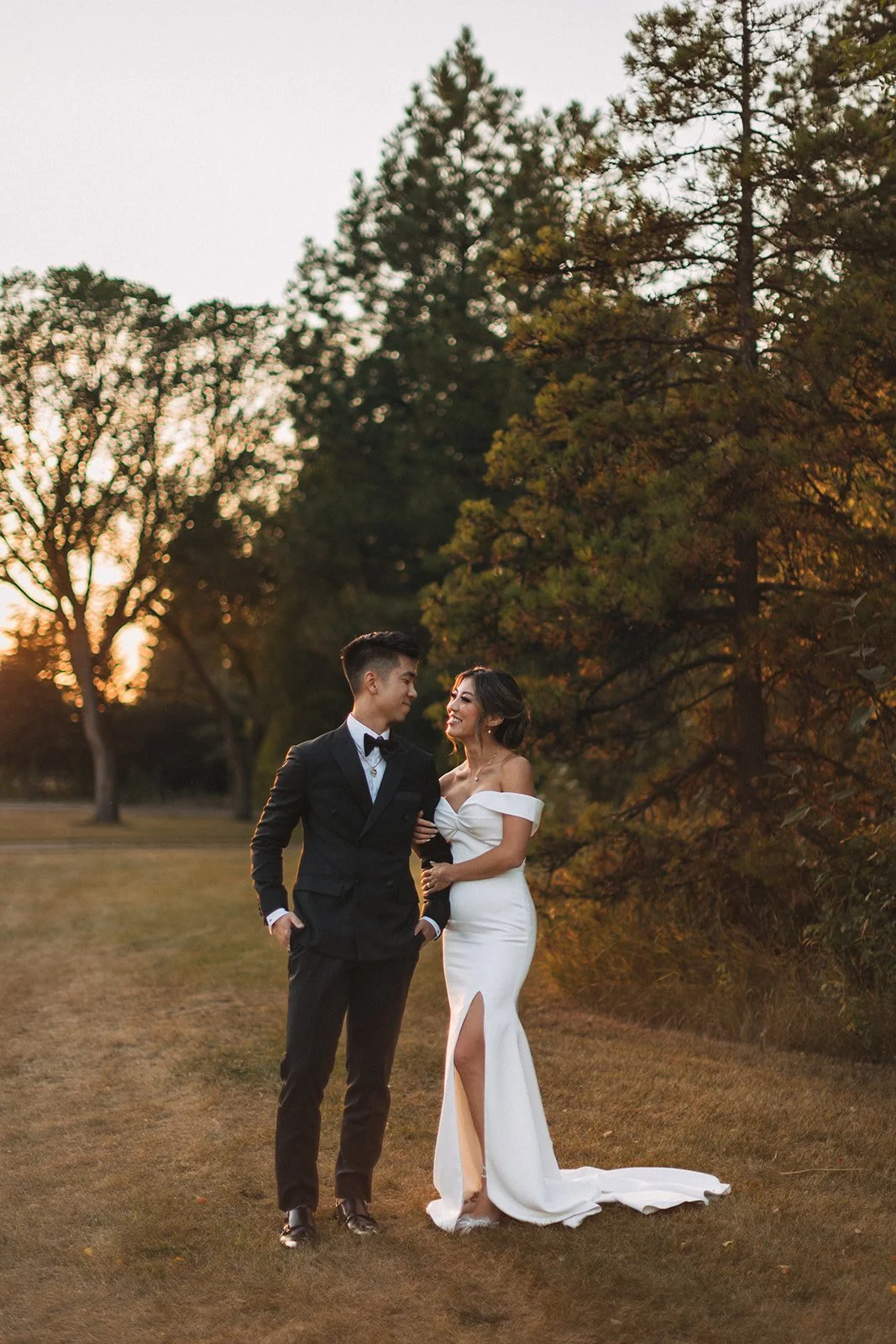 A couple steps out of their reception for sunset photos, posing and looking at each other while the sun sets against the trees behind them