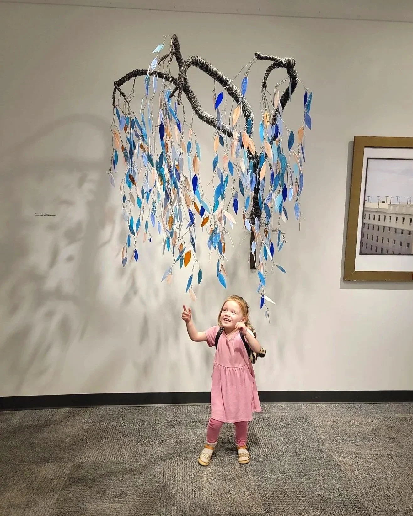 A young girl in a pink dress and leggings standing in front of a wall with a hanging art installation designed to look like a tree with blue, beige, and orange leaves made of paper or fabric. She is smiling and pointing upward.