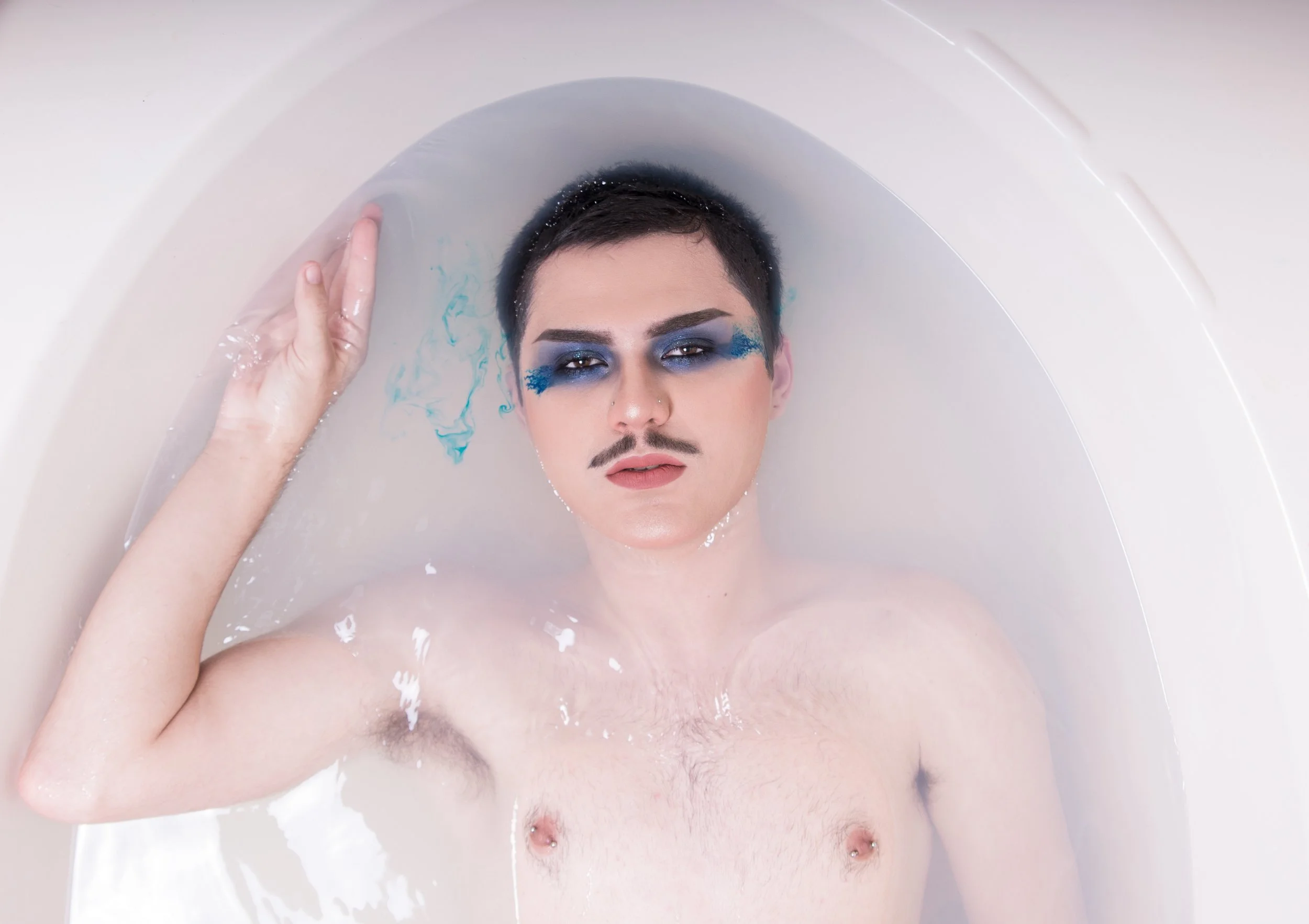 Person with makeup and a fake mustache lying in a bathtub filled with water, with one hand resting on the tub edge.