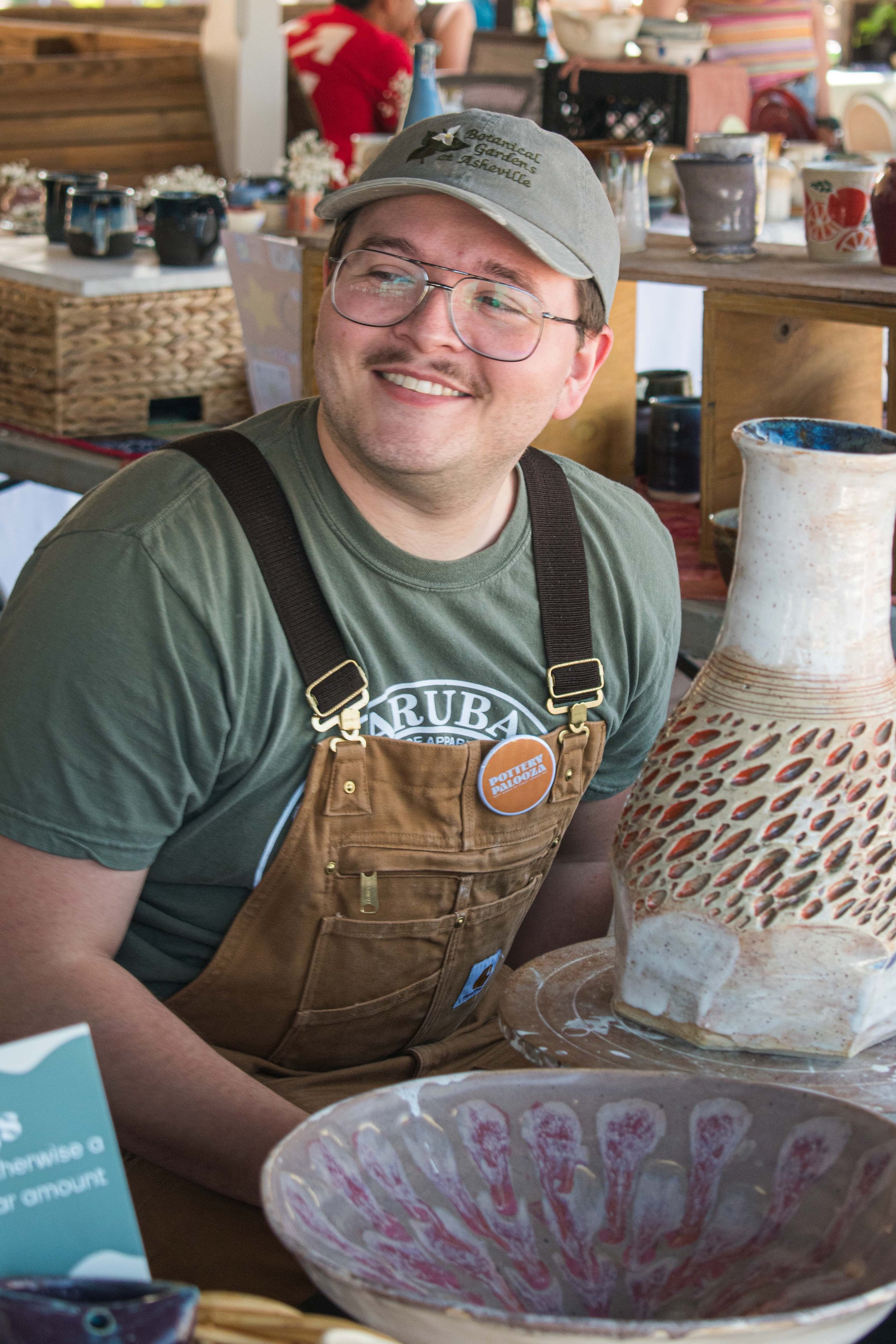 A smiling man in glasses, a green T-shirt, and brown apron sitting at a pottery booth with ceramics and pottery pieces around him.