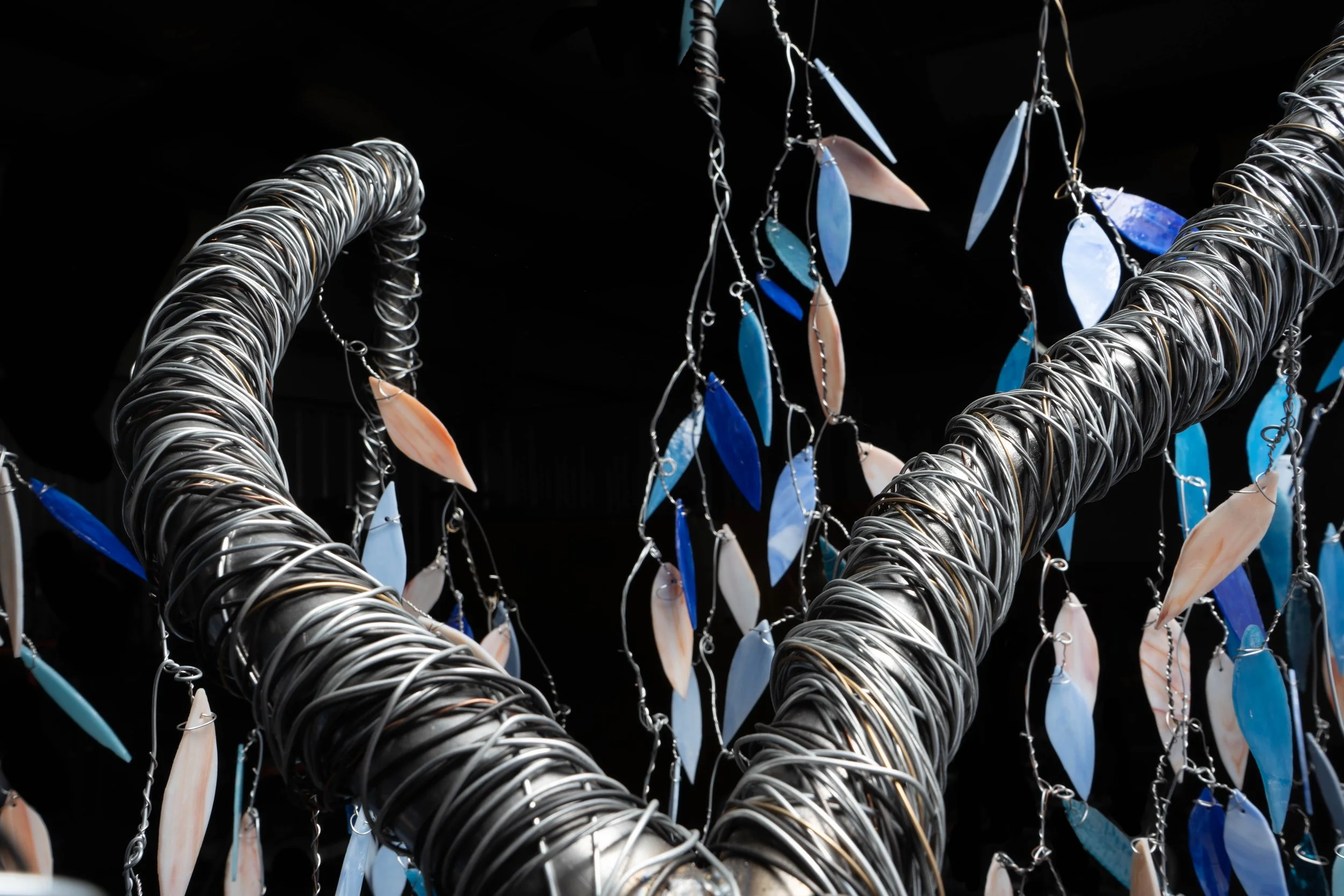 Close-up of a handcrafted jewelry display featuring metal wire sculptures with hanging shells or stones in shades of blue, white, and peach, set against a dark background.