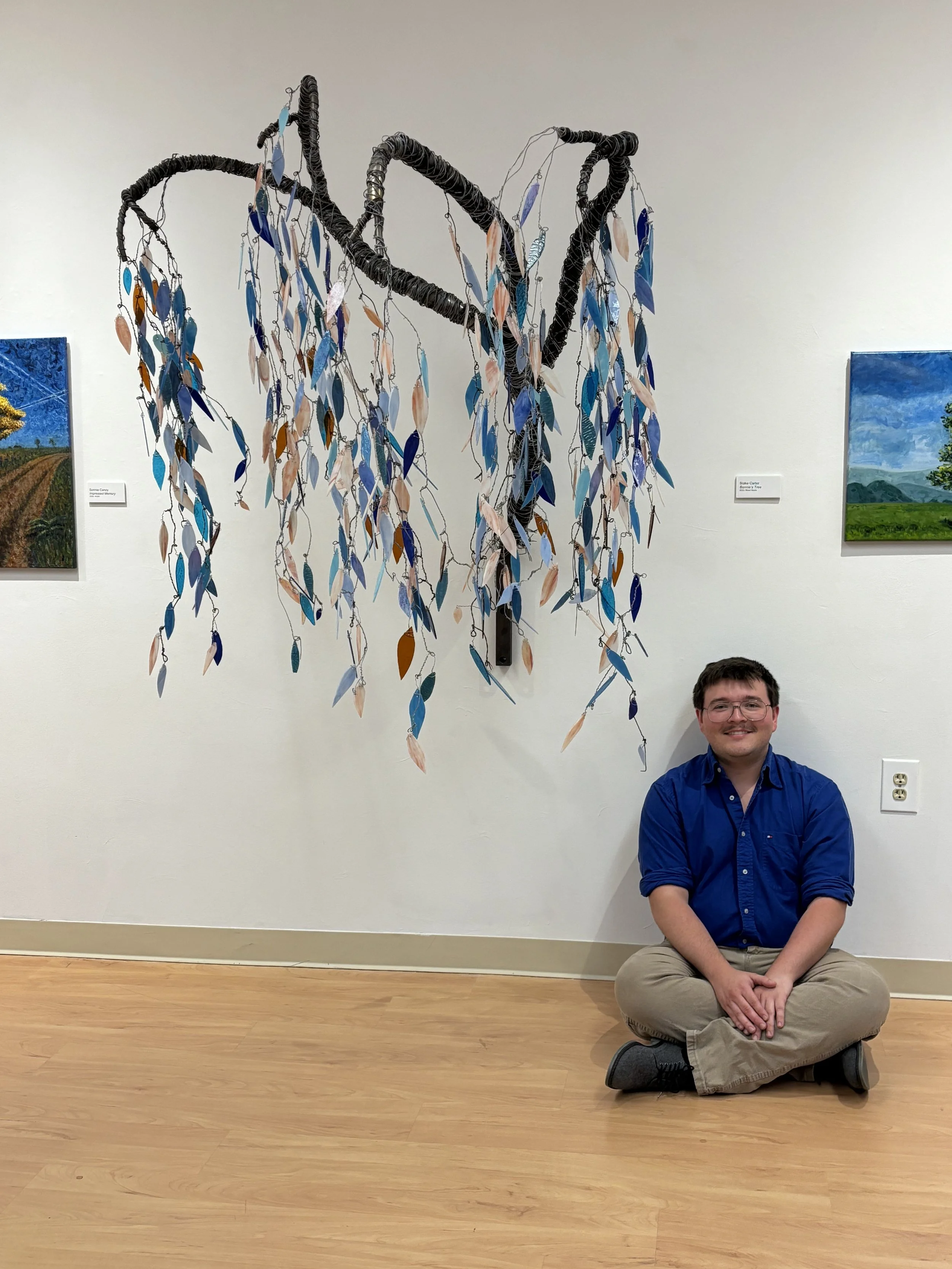 A person sitting cross-legged on the floor in front of a wall-mounted art installation that resembles a tree with hanging leaves. The person is wearing a blue shirt and khaki pants, smiling at the camera. The art installation is made of black materials with paper leaves in various shades of blue, beige, and brown hanging from wire.