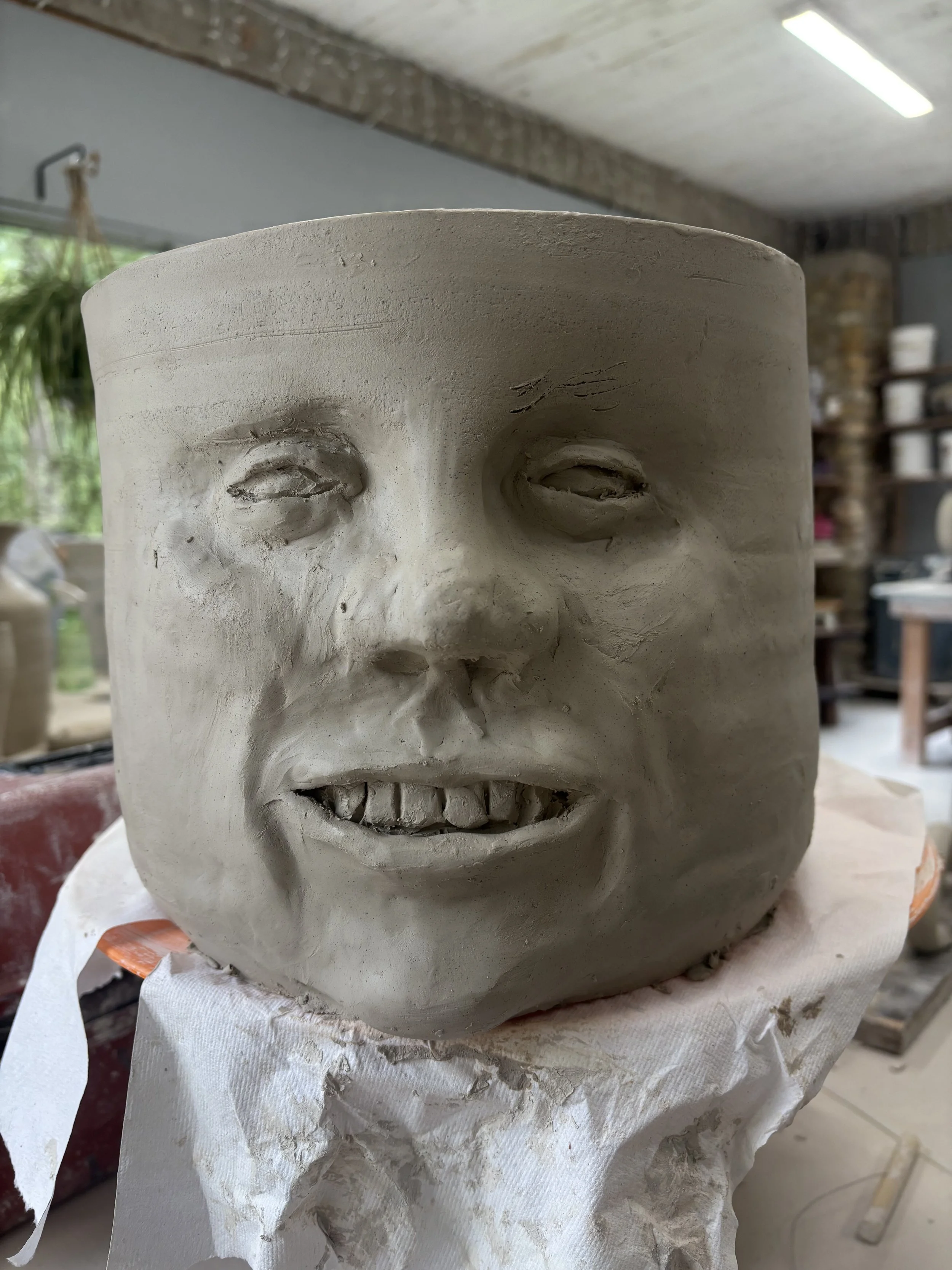A ceramic sculpture of a human face in progress, featuring detailed facial features on a large, cylindrical form in an artist's studio.