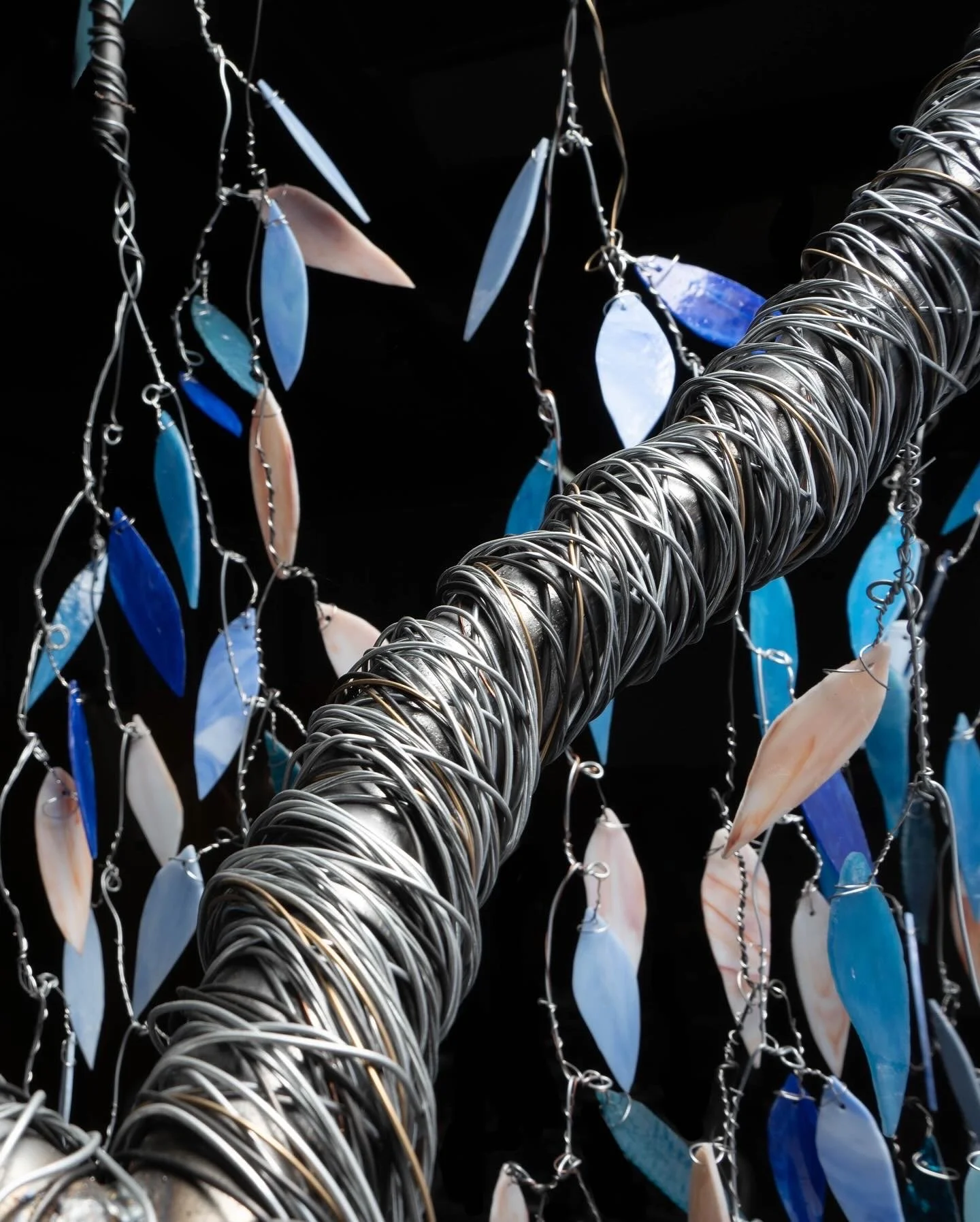 Close-up of a sculpture made of intertwined metal wires with hanging blue and beige shell-like pieces against a black background.