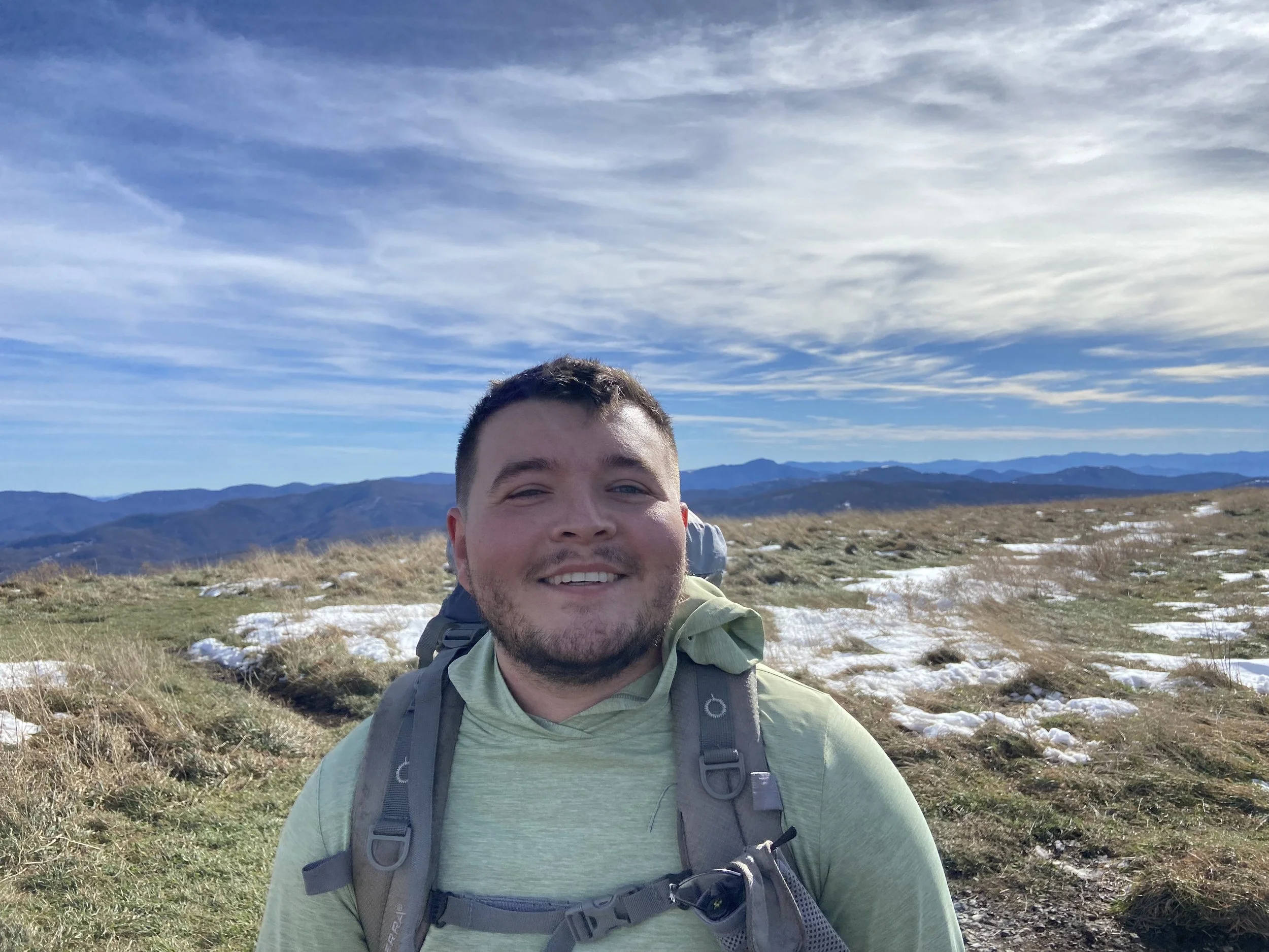 A smiling man with a backpack standing outdoors on a mountain trail with rolling hills and snow patches, under a partly cloudy sky.