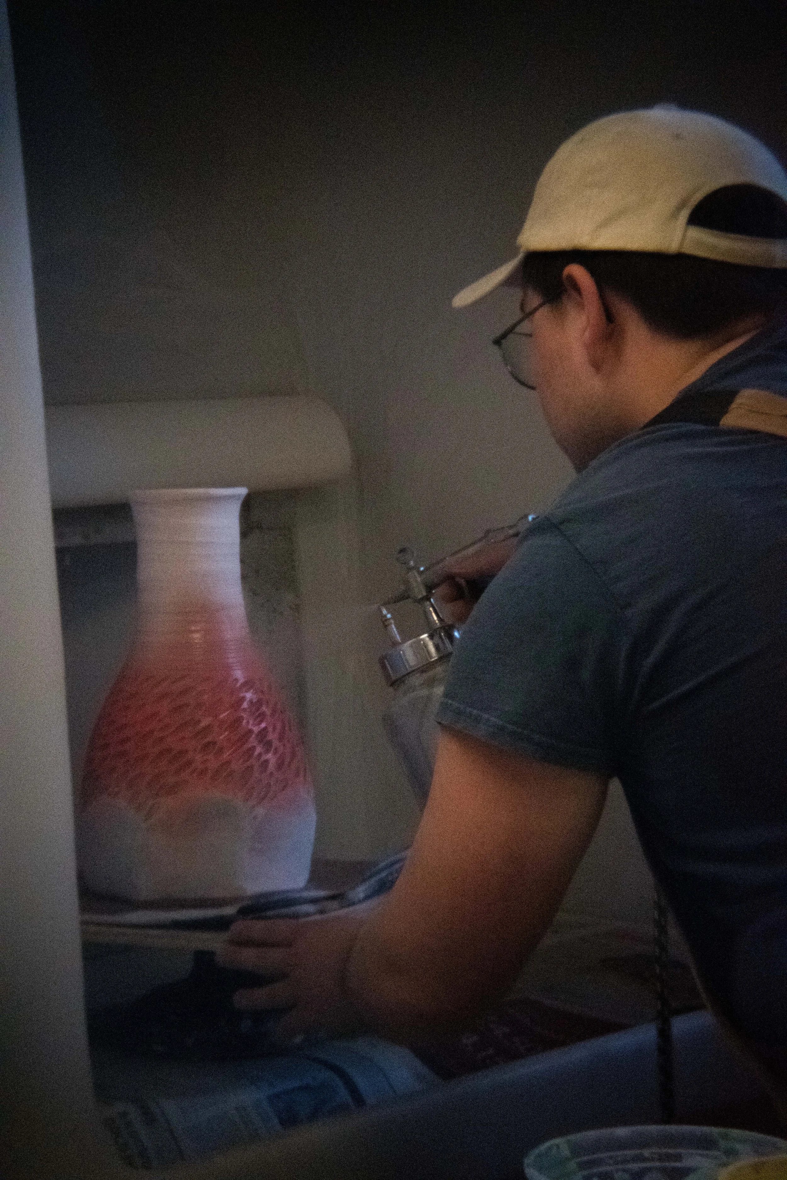 A person wearing a white cap and glasses is spray-painting a vase with pink and white designs inside a dimly lit space.