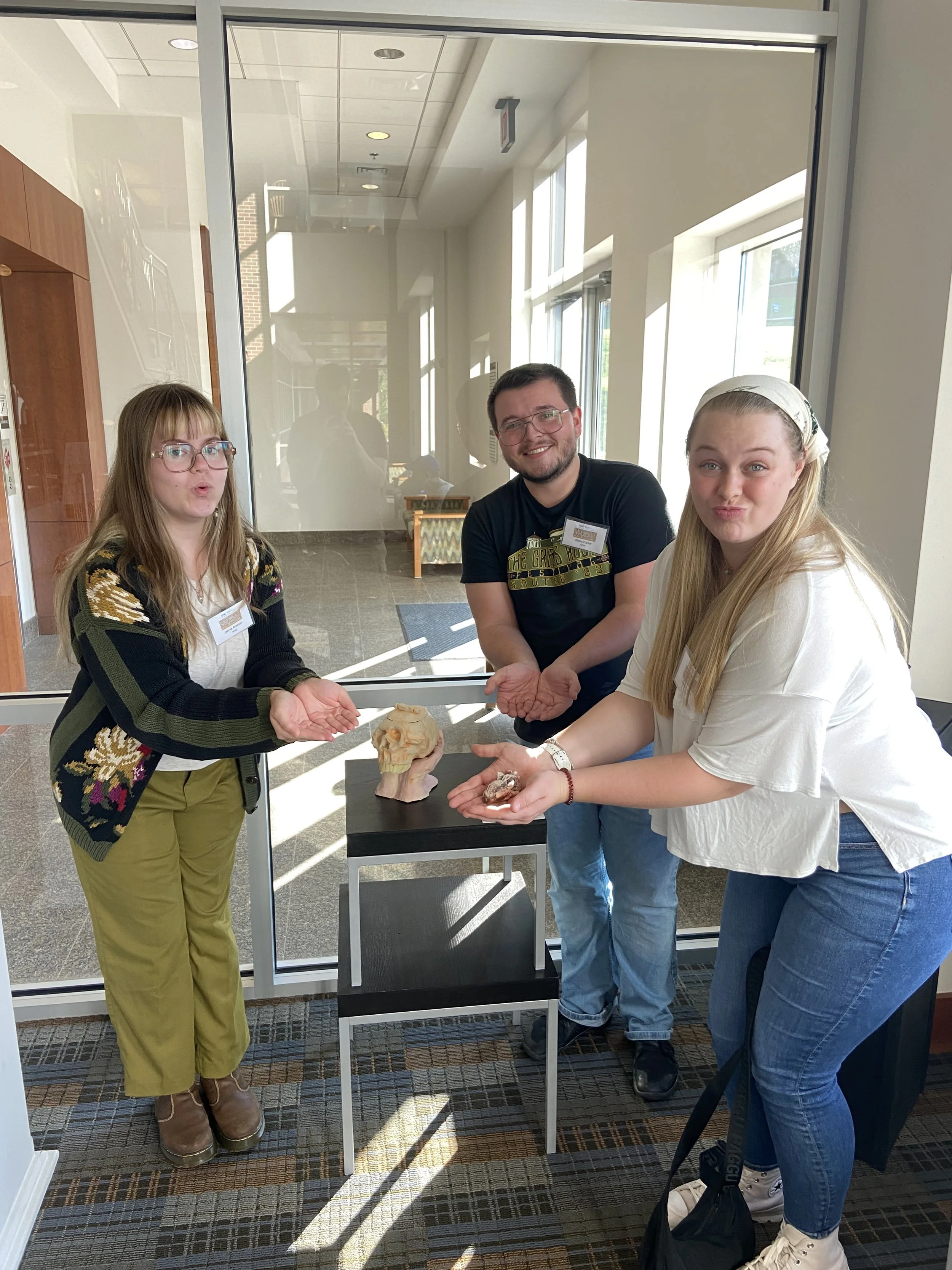 Three people, two women and one man, holding rocks and minerals inside a building near a large window, with a skull model on a small table between them.