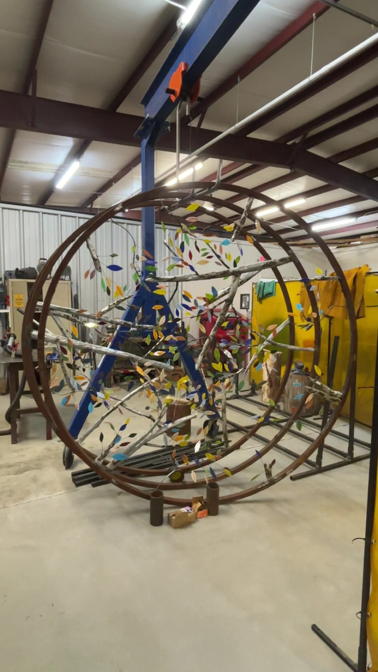 A workshop with a large metal sculpture resembling a tree with colorful leaves inside a circular frame, with tools and equipment around.