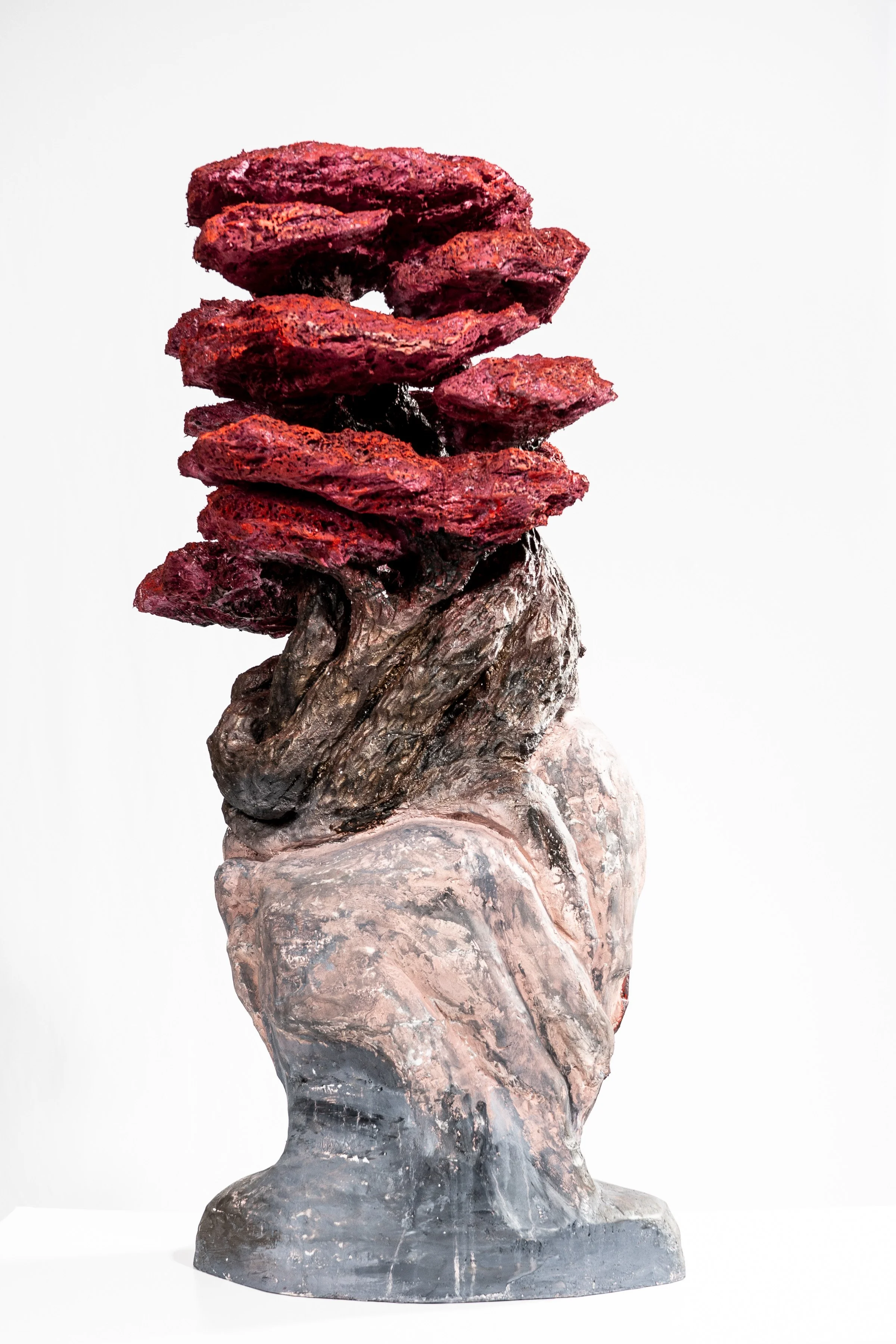 Sculpture of a rough-textured head with a stack of red, rocky shapes on top, resembling hair, against a plain white background.