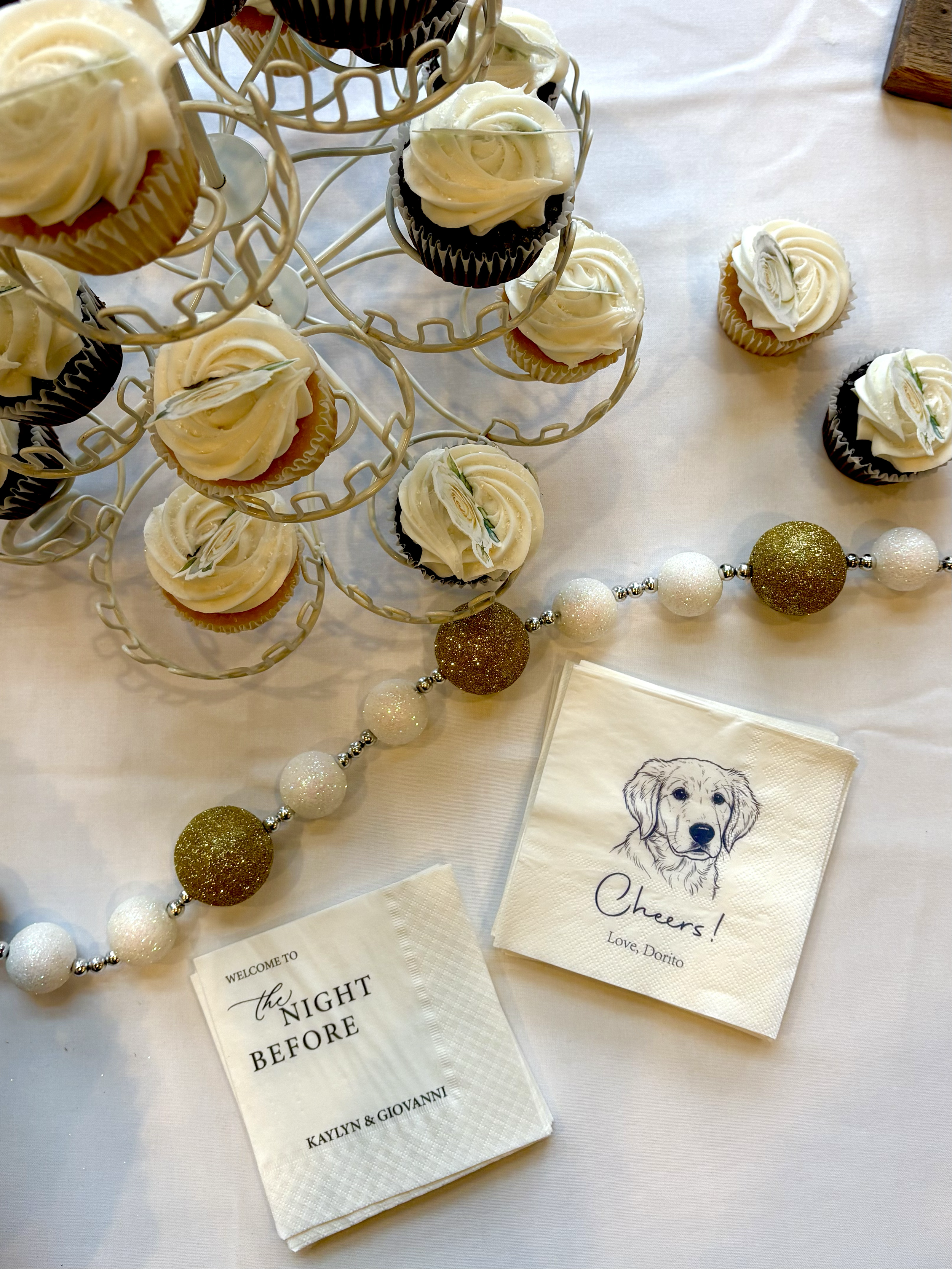 Cupcakes with white frosting and gold and black decoration on a cupcake stand, surrounded by white and gold glittery bead decorations, and paper napkins with dog illustrations and messages for an event.