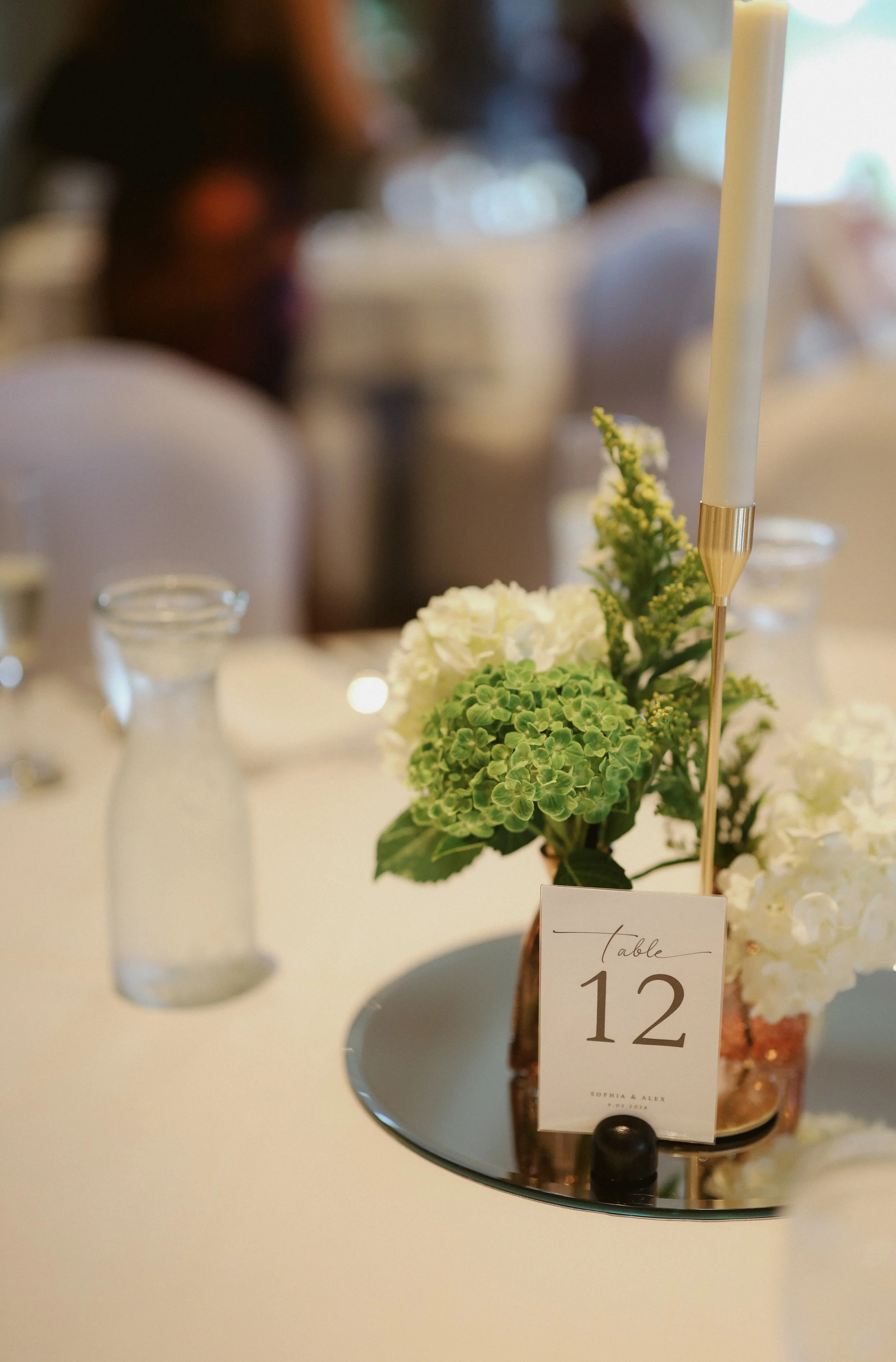 A floral centerpiece with green hydrangeas, white flowers, and a tall white candle on a table with a mirrored surface, labeled with a table number card reading 'Table 12.' The background is blurred with a person visible.