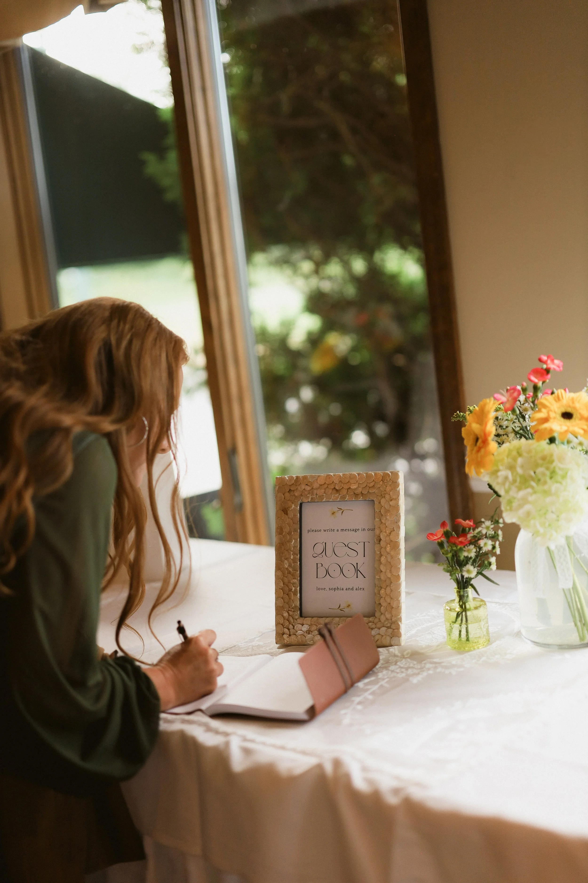 A woman with long wavy hair writing in a guest book at a table decorated with flowers, with a sign that reads 'Guest Book' and a message for guests to write in.