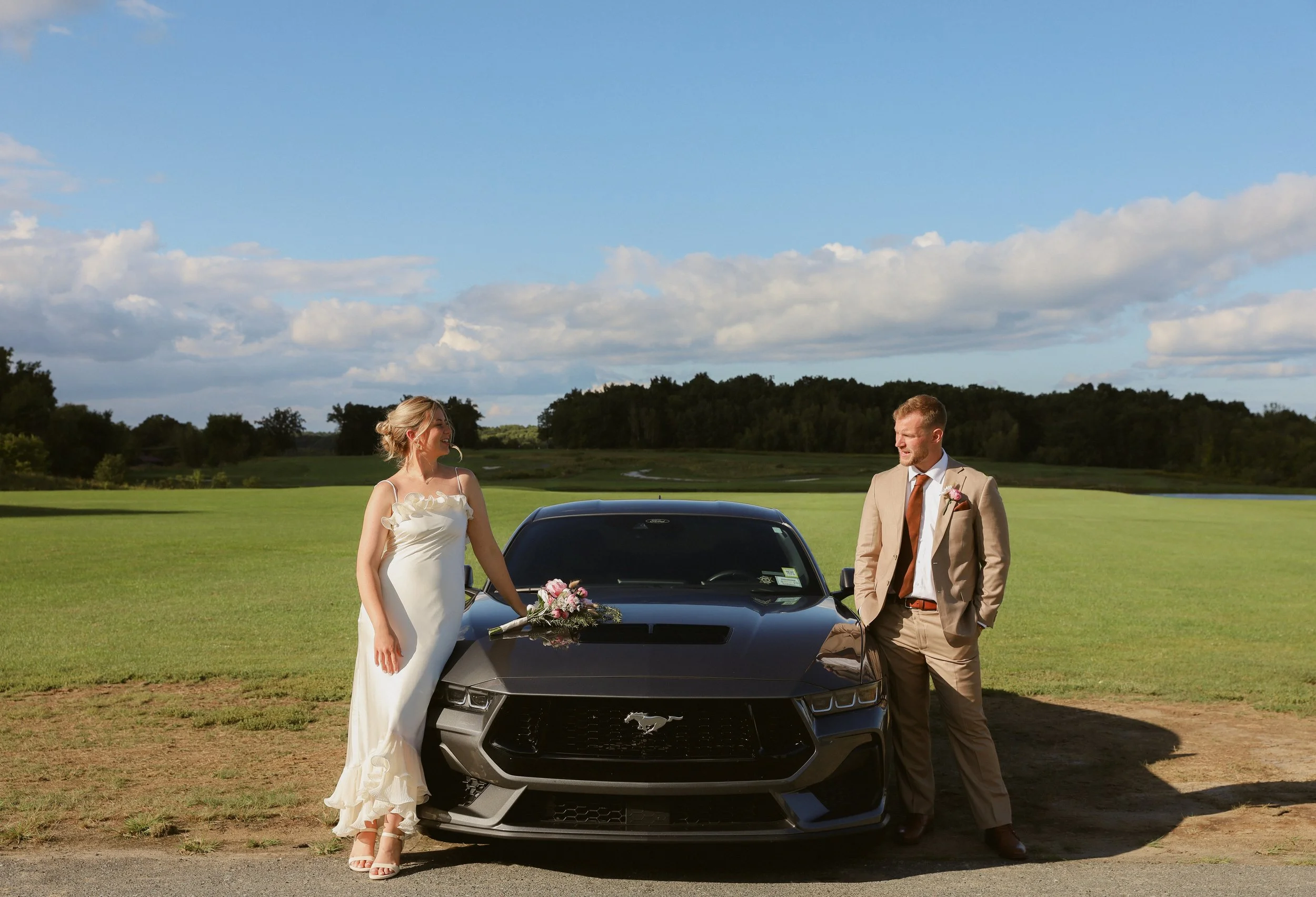 A bride and groom standing outside near a black Ford Mustang, with a field and trees in the background under a partly cloudy sky.