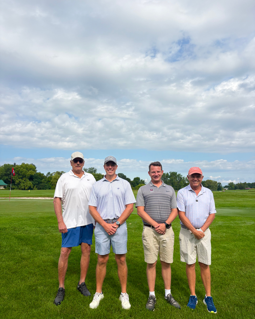 Four men standing on a golf course with green grass, trees, and a cloudy sky in the background. They are dressed in golf attire and smiling at the camera.