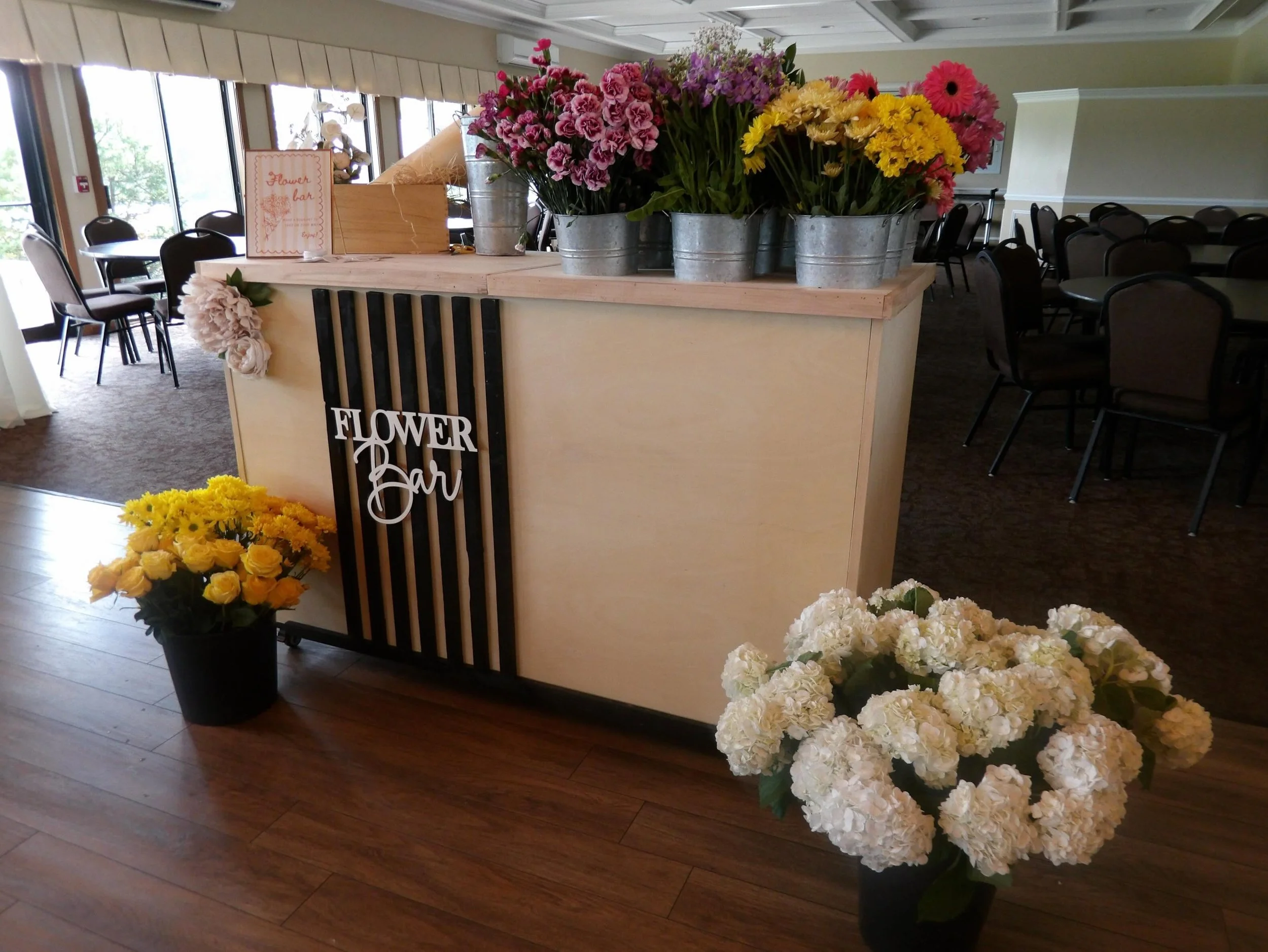 A flower bar stand decorated with pink, purple, yellow, and white flowers in metal buckets. The stand has a sign that reads "Flower Bar" and is in a room with chairs and tables, possibly set up for an event or wedding.