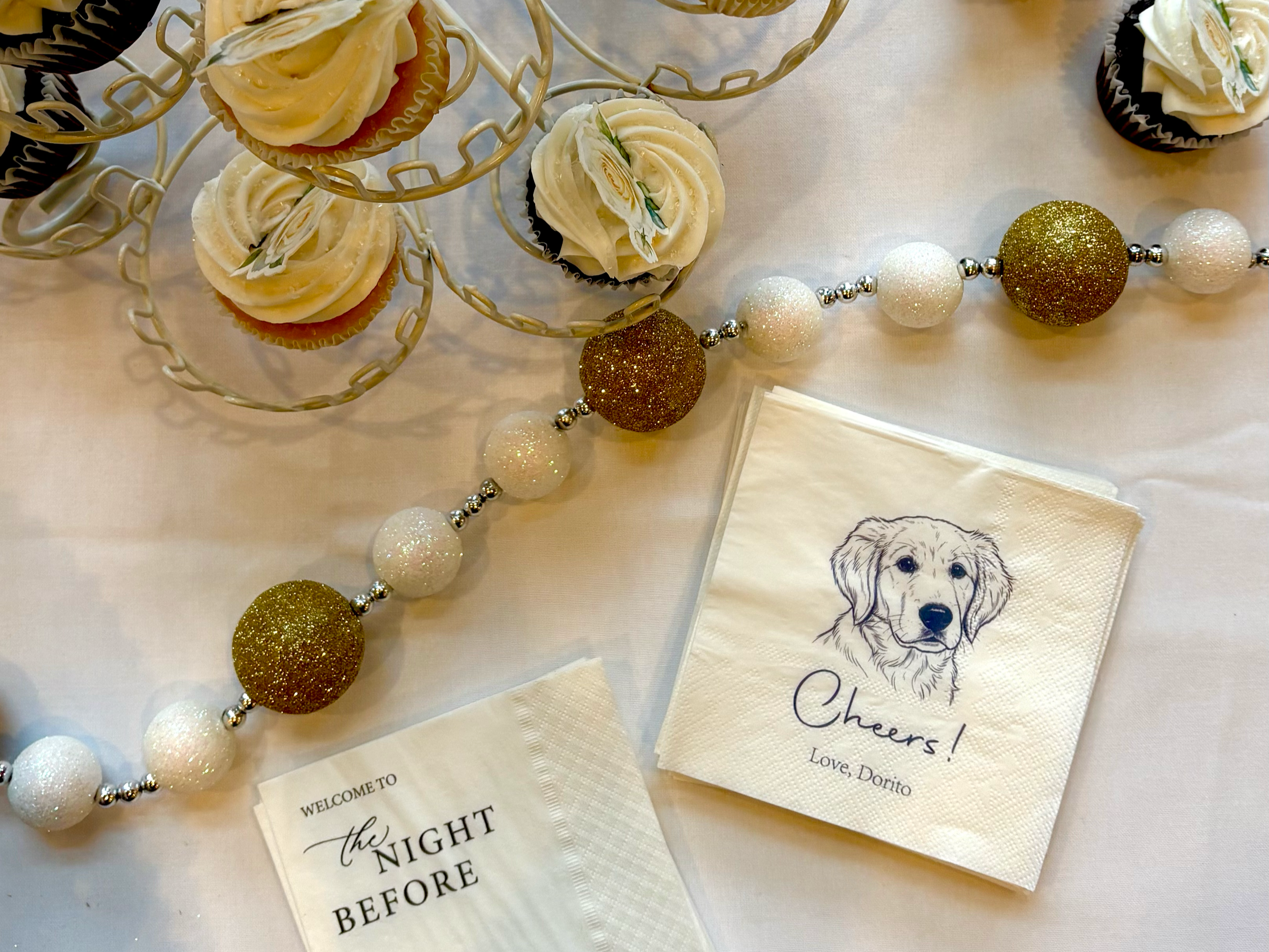 Decorative cupcakes with white frosting and small paper toppers featuring a dog's face, set on a white table with festive gold and white baubles and greeting cards.